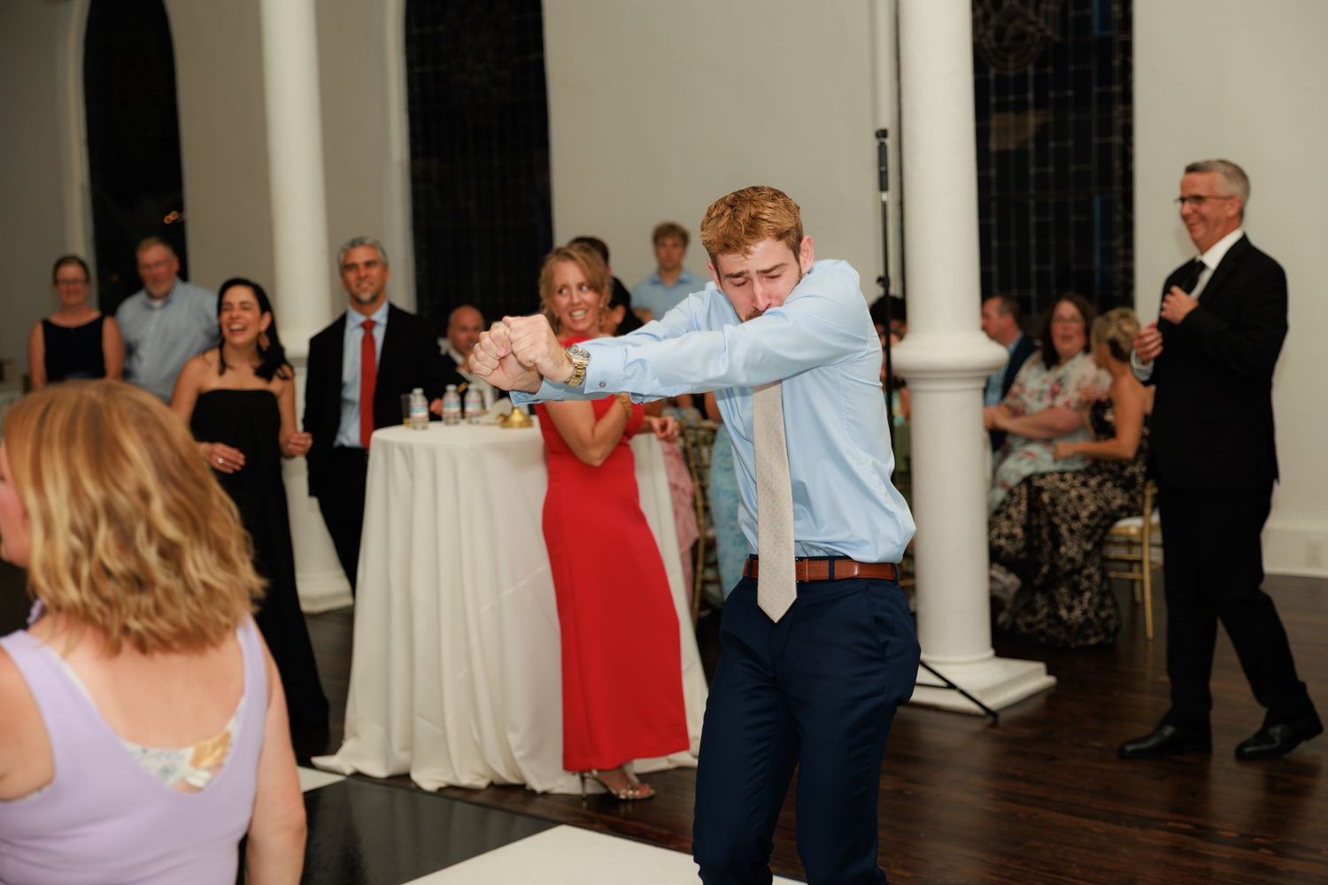 Young man dancing energetically at wedding reception while guests watch and smile.