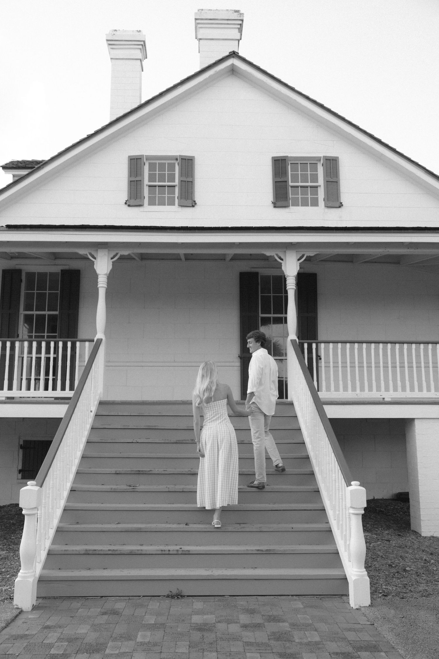 Black and white photo of a couple holding hands walking up stairs to a classic white colonial house with dark shutters.