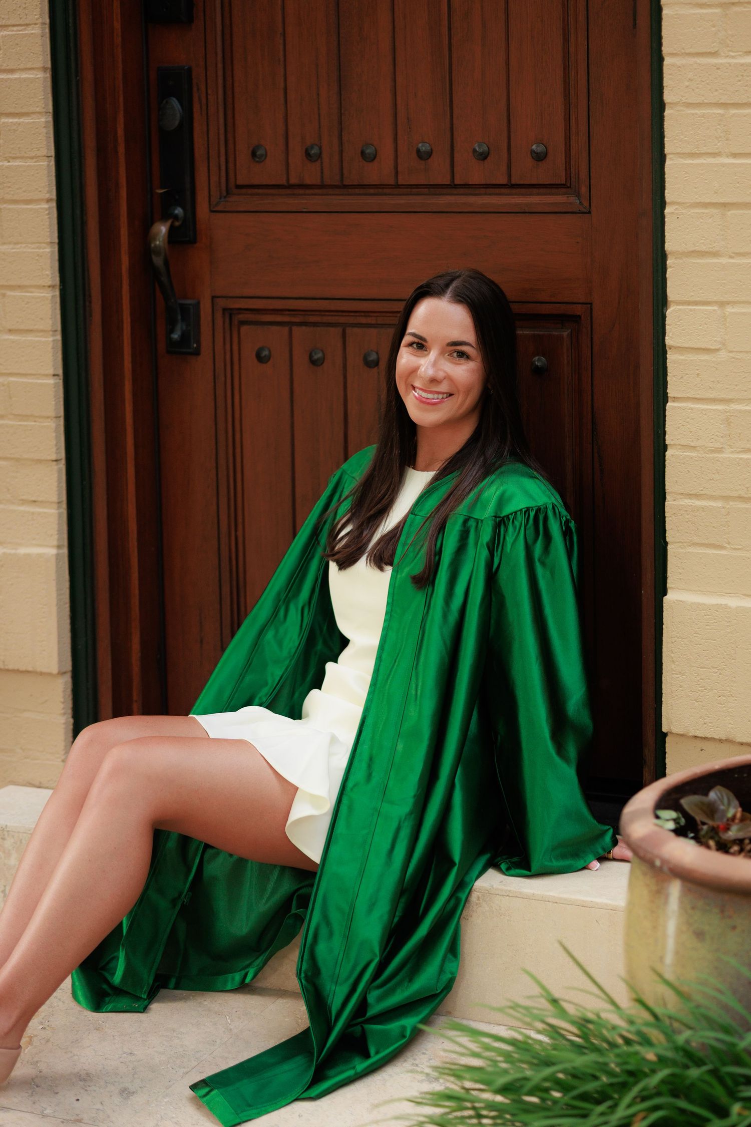 Young woman in green graduation gown sits smiling on steps in front of a wooden door wearing a white dress.