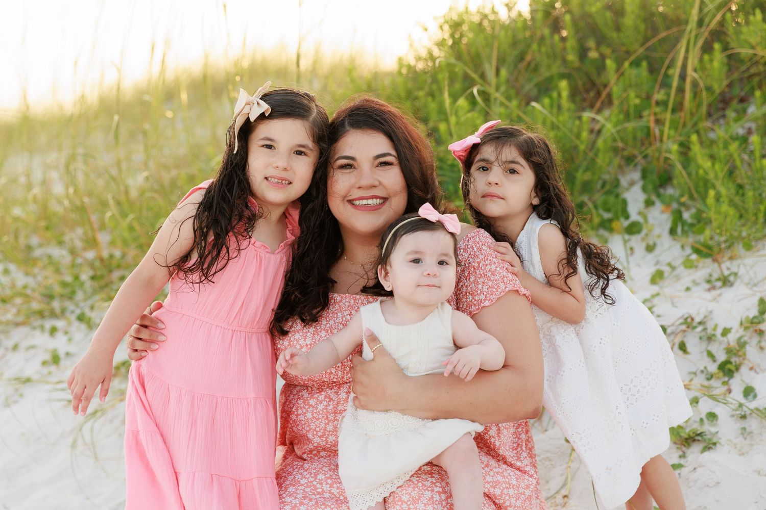 A smiling mother in a floral dress holds her baby at the beach, surrounded by two young daughters in pink dresses.