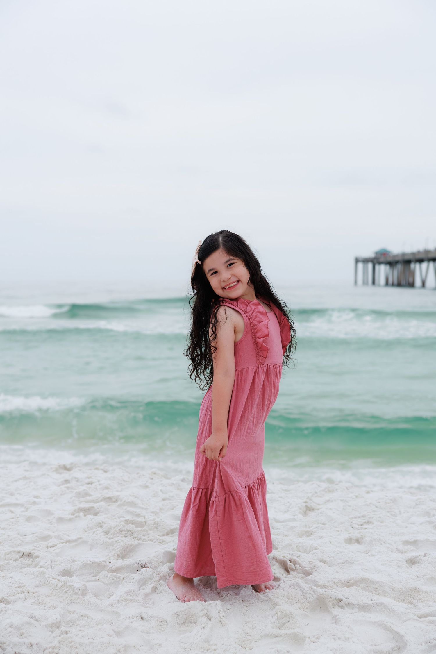 A young girl in a pink maxi dress smiles on a white sandy beach with turquoise ocean waves behind her.