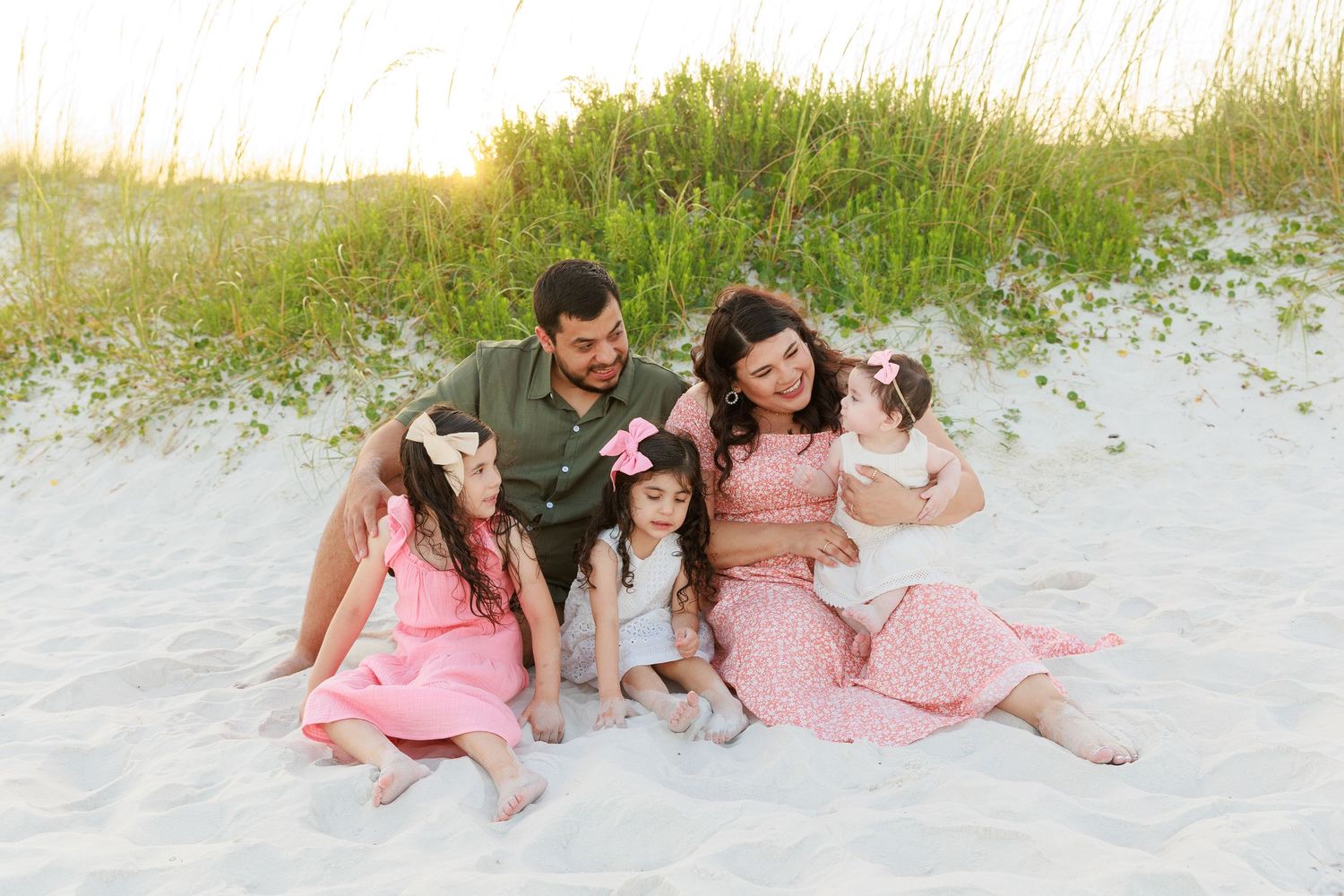 Family of five sitting on white sand beach, parents and three young girls in pink dresses smiling together near grass dunes.