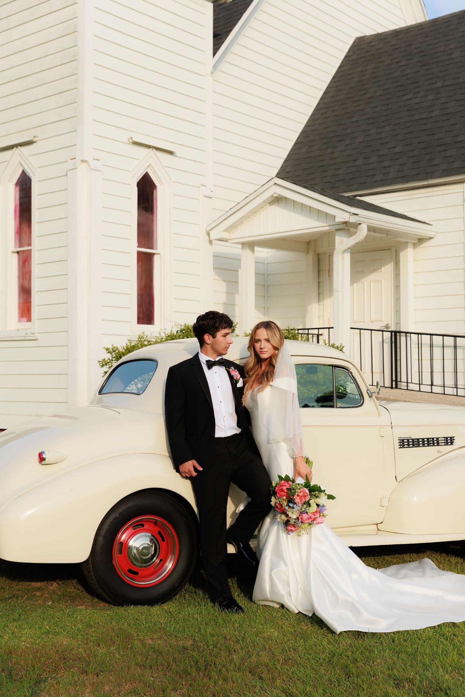 A couple poses by a vintage cream-colored car outside a classic white church with red-trimmed windows.