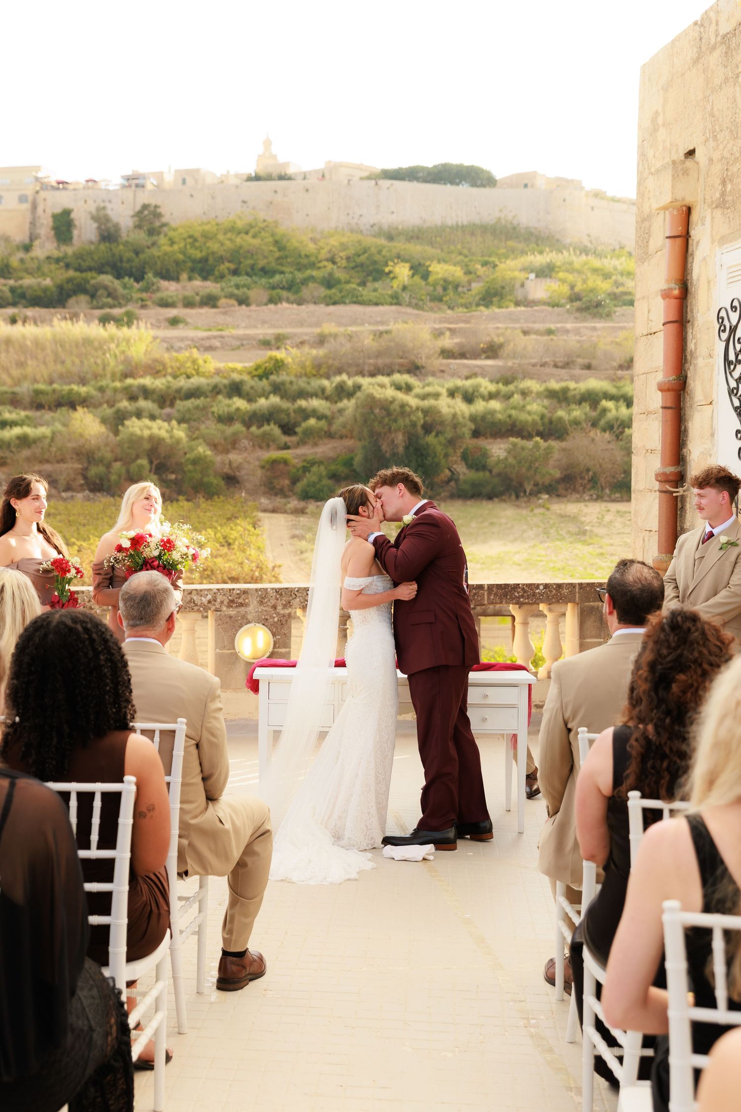 Outdoor wedding ceremony with guests seated in white chairs while couple exchanges vows under rustic pillars.