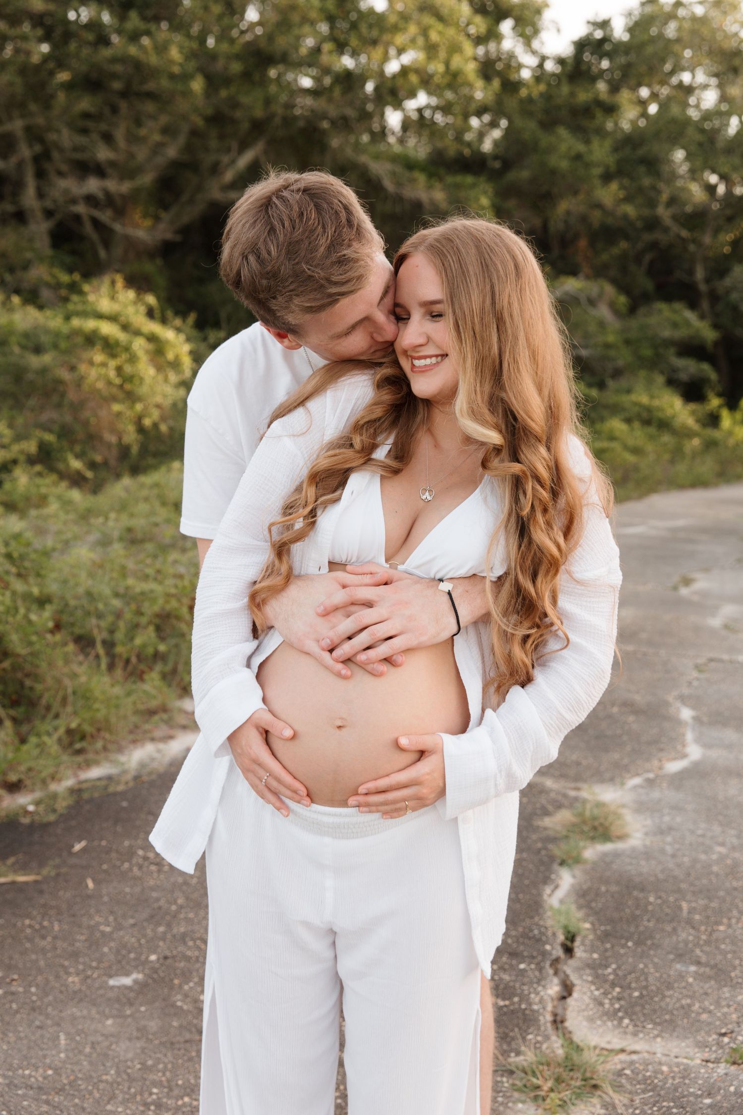 Maternity photos showing couple in white clothing embracing on outdoor pathway.