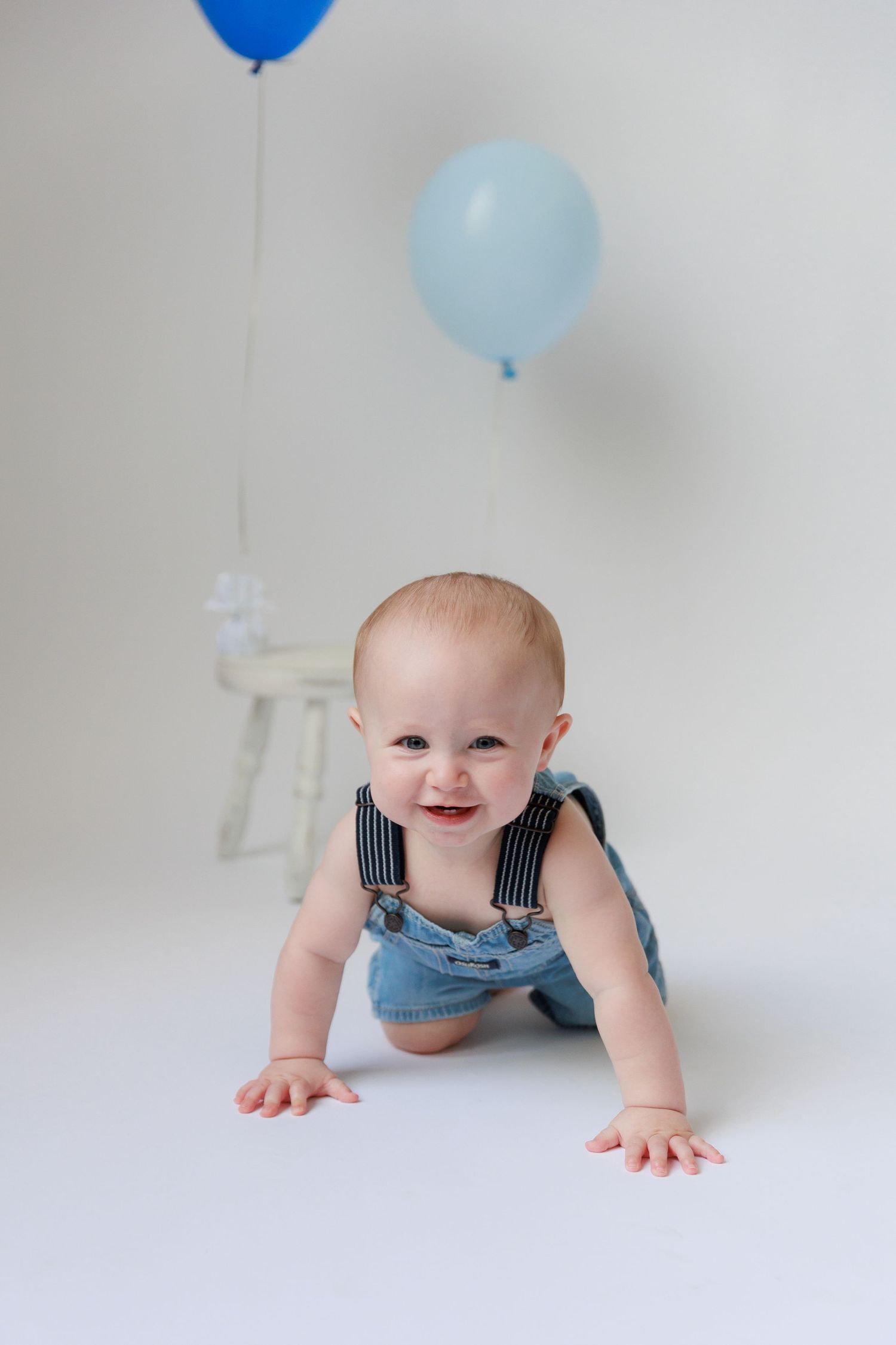 Baby in denim overalls crawls on white floor with blue balloons floating overhead in studio setting.