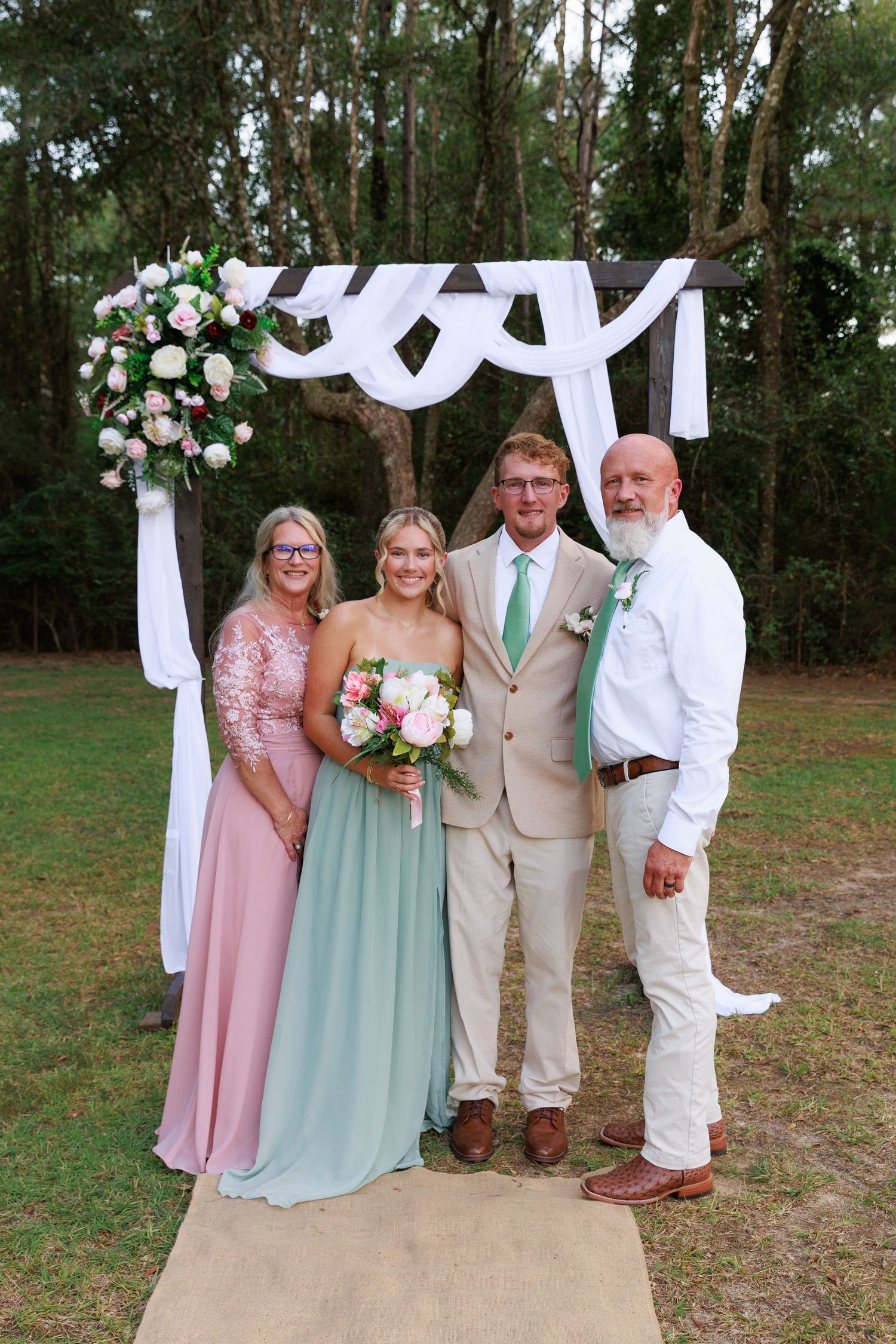 Wedding party poses in front of white draped arch outdoors with pink and mint green attire.