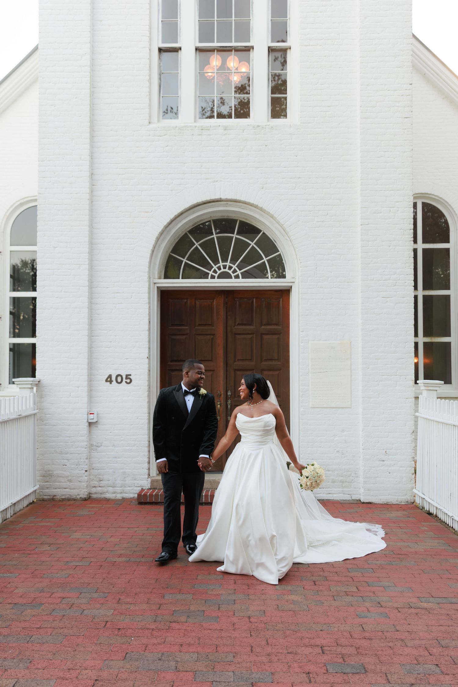 A wedding couple poses in front of a classic white church entrance with red brick steps.