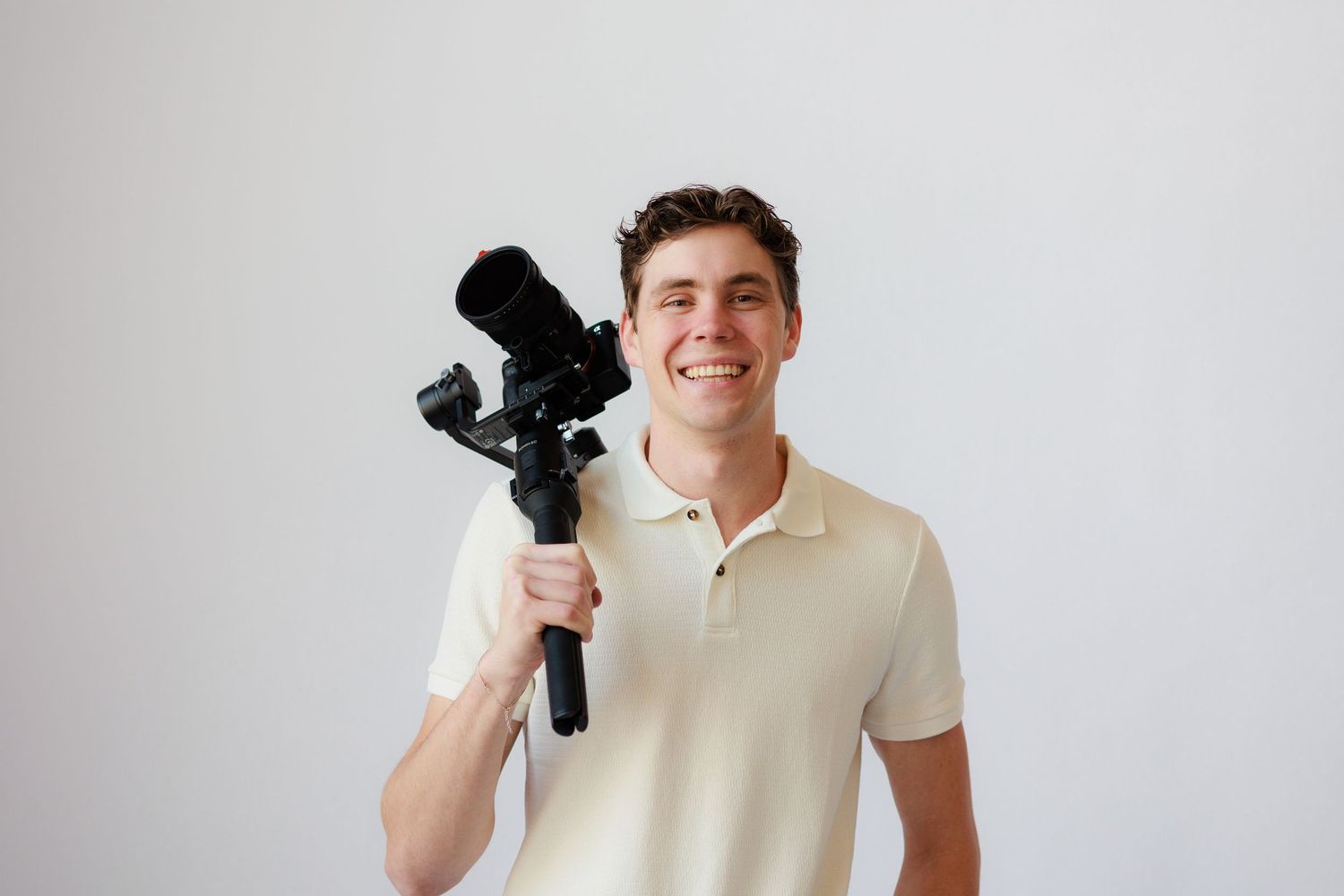 Professional photographer wearing cream polo shirt holds camera gimbal while smiling against white background.