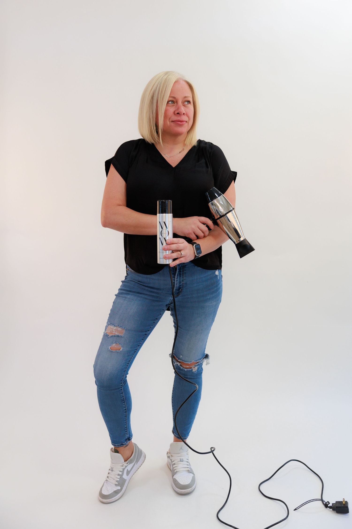 Stylist holding professional hair tools while posing in casual outfit against white background.