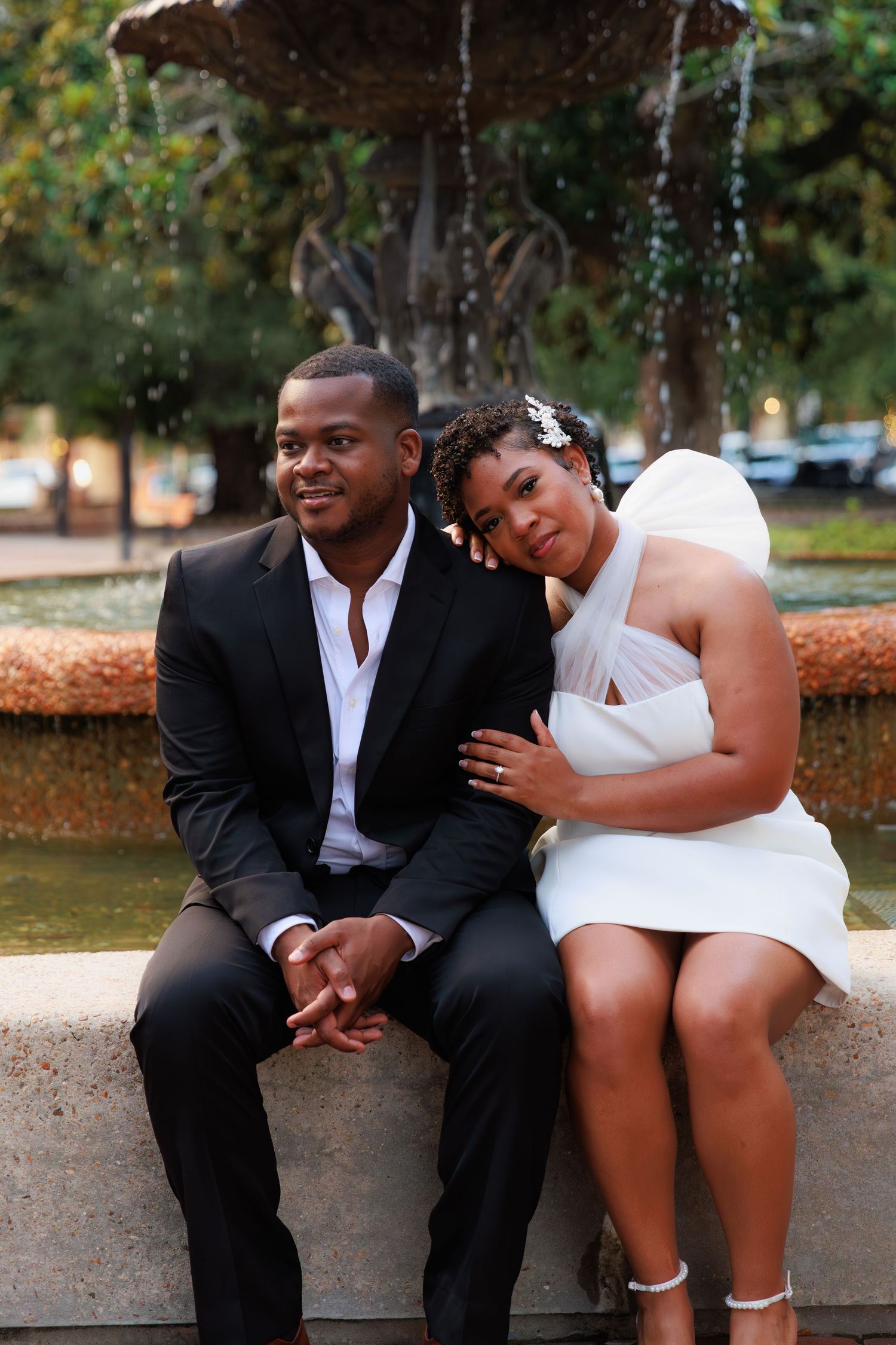 Bride and groom sitting together on a fountain ledge, smiling, bride in white mini dress and groom in black suit.