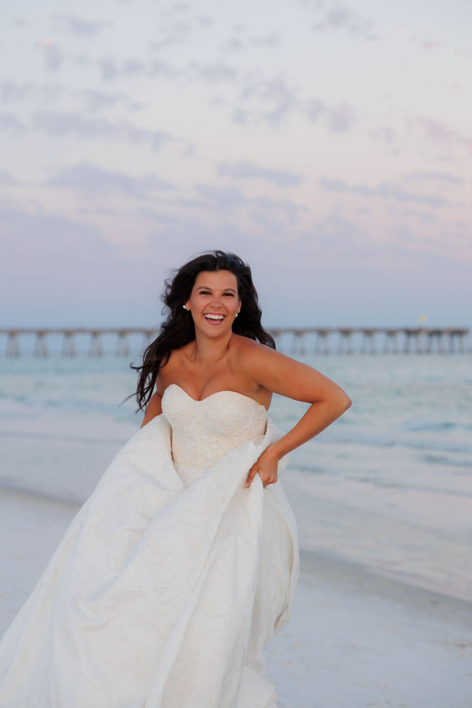 Joyful bride in strapless white wedding gown laughs on beach at sunset with pier visible behind her.