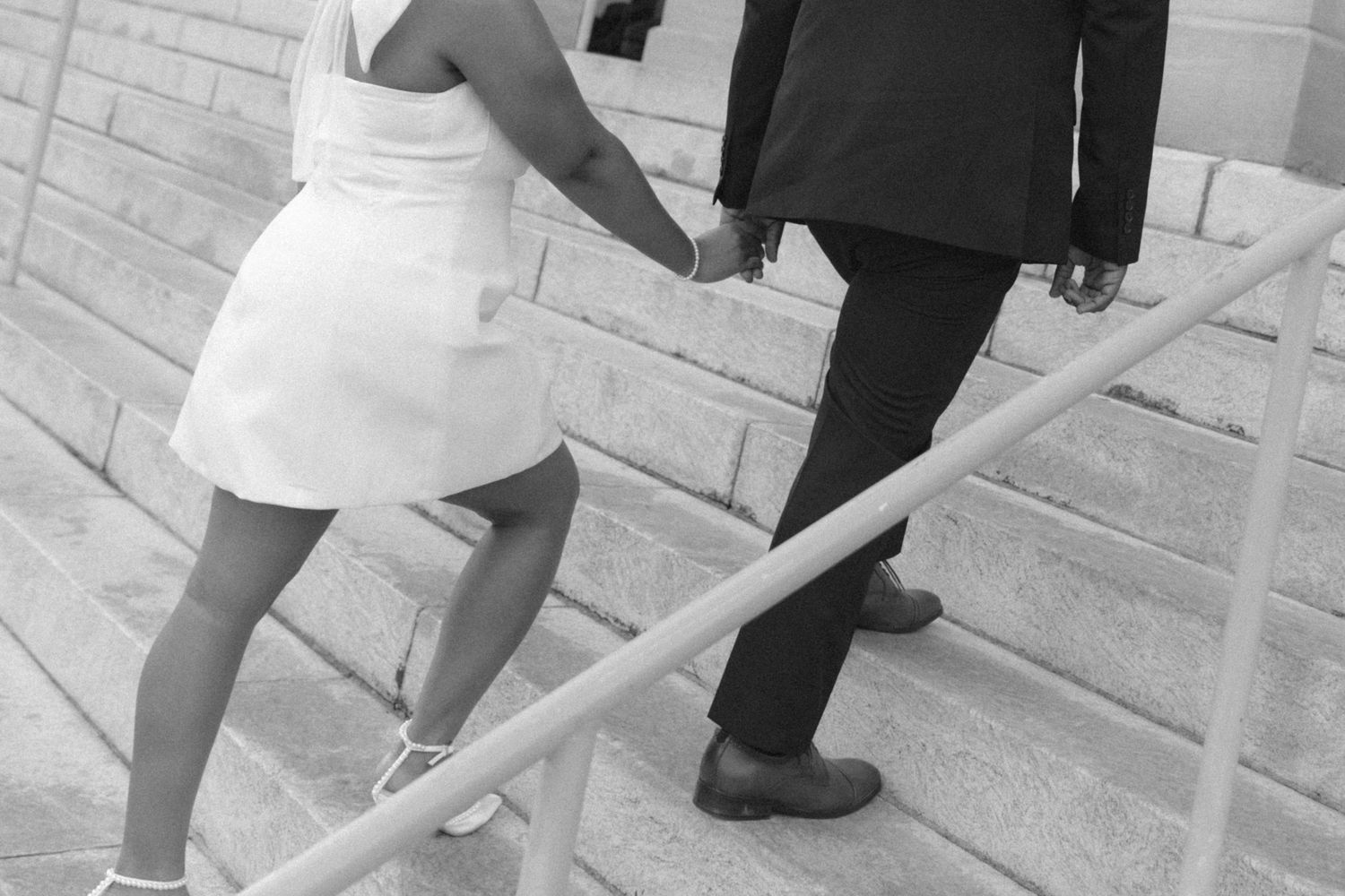 Black and white photo of a couple holding hands walking up stone steps, woman in white dress and man in dark suit.