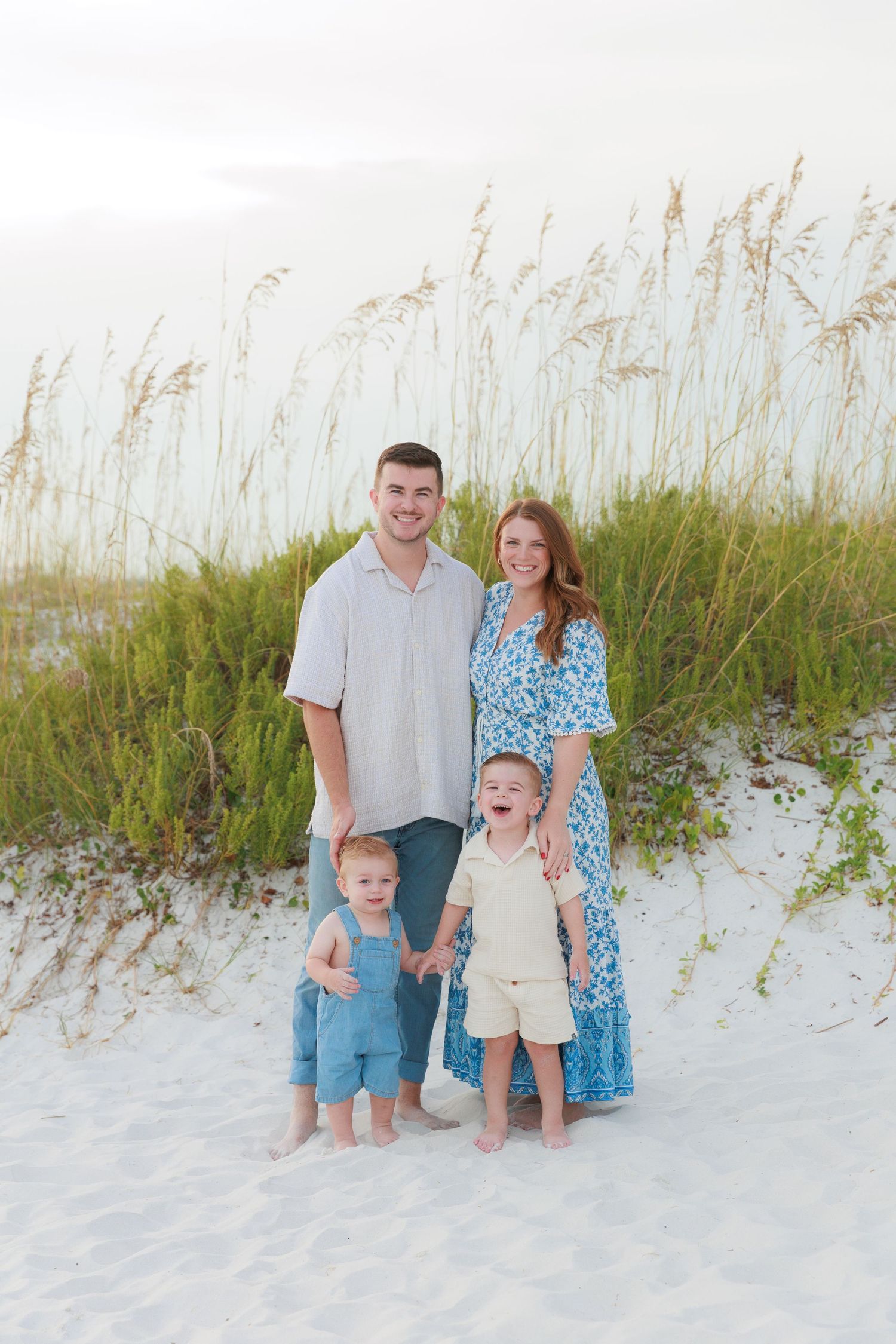 Young family of four smiling together on white sand beach with sea oats and dunes behind them.