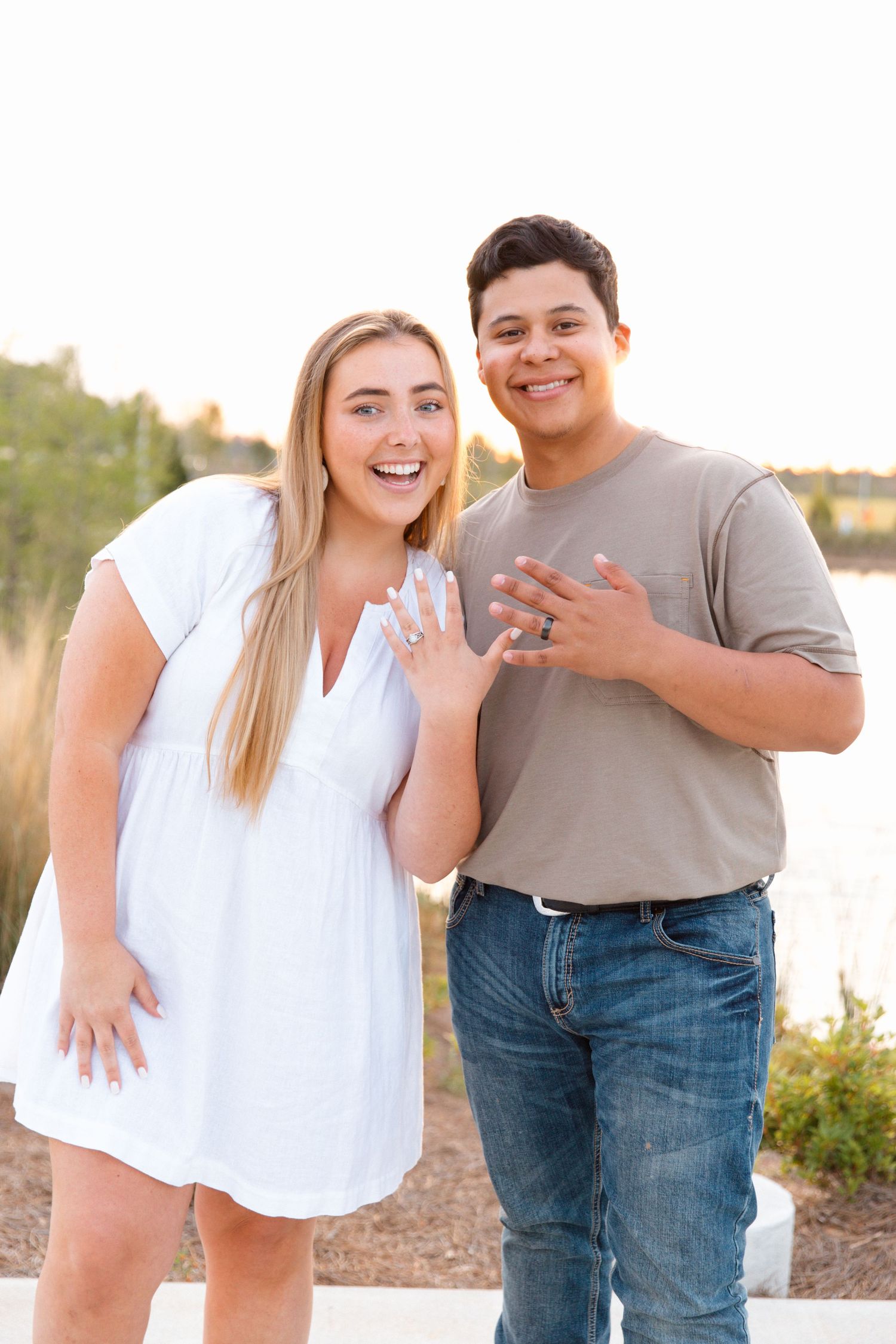 Happy couple showing off engagement rings outdoors near a lake at golden hour.