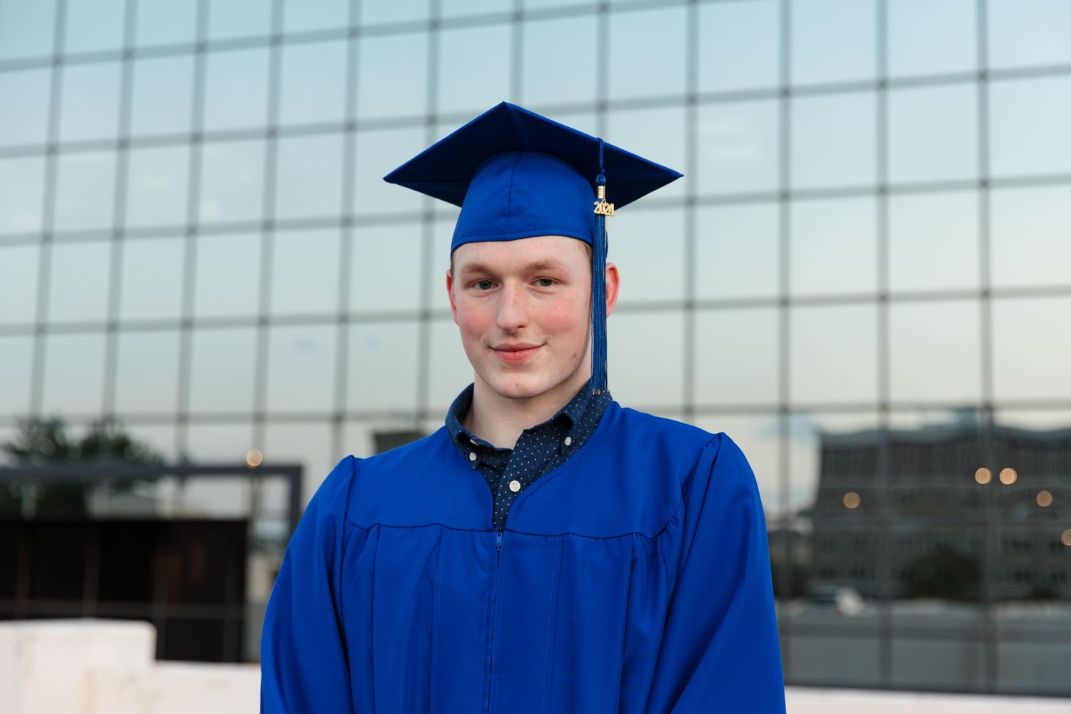 Young man in blue graduation cap and gown smiling in front of glass building.