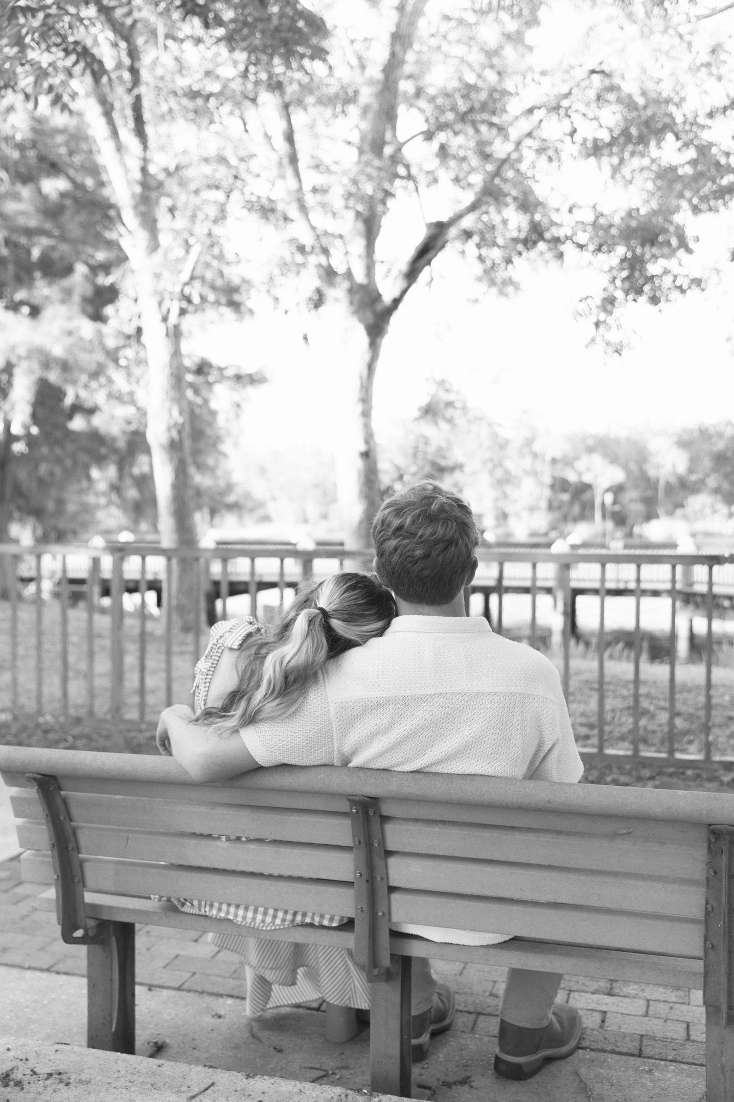Black and white photo of a couple sitting close on a park bench, viewed from behind, surrounded by trees.