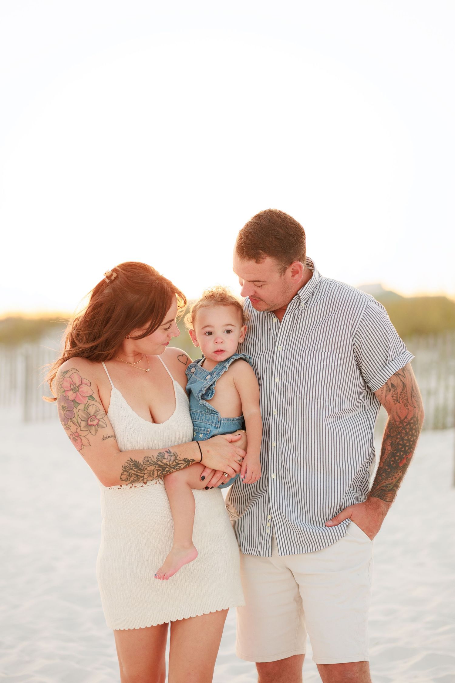 Tattooed parents in casual attire lovingly gazing at their baby on a bright sandy beach.