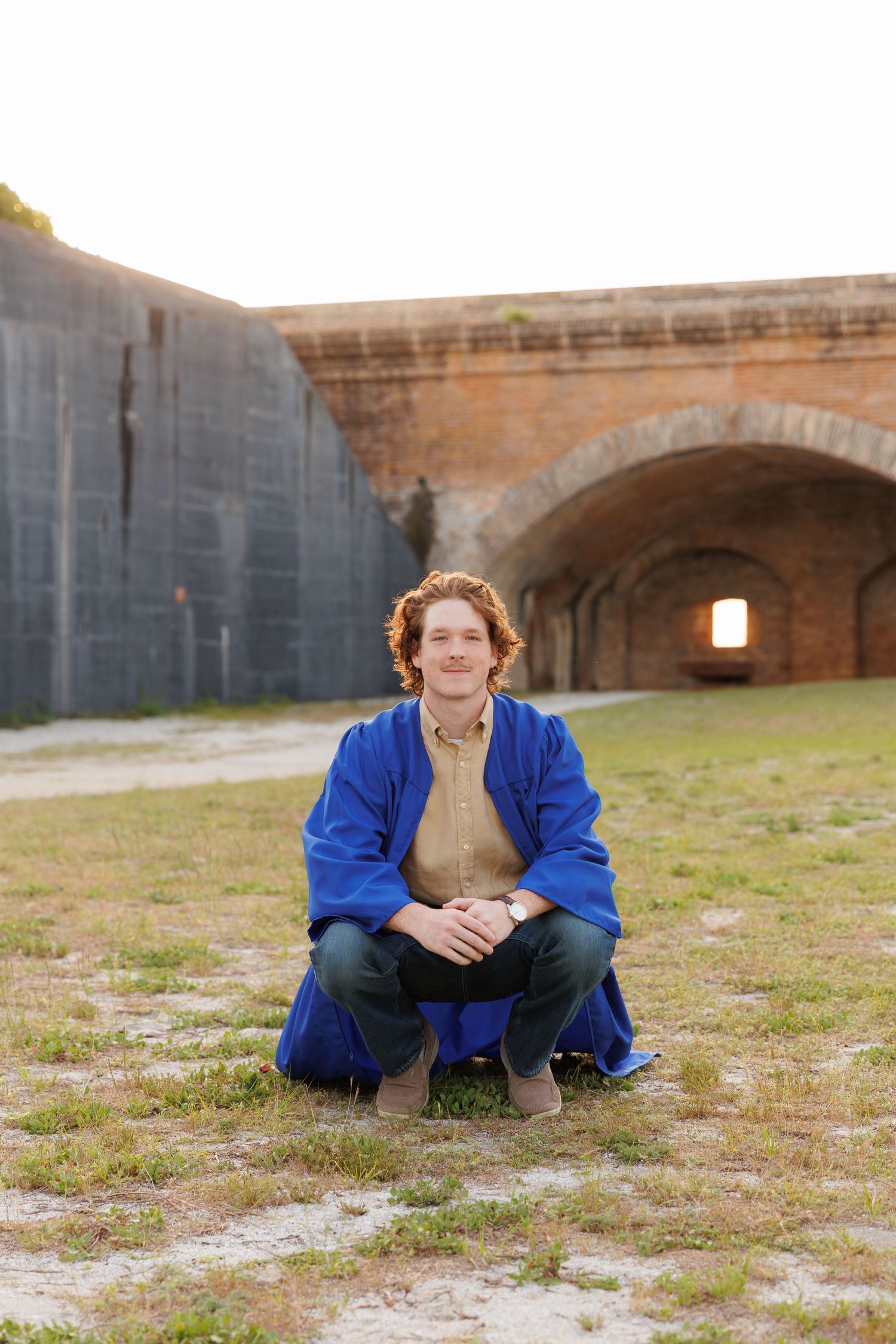 Young man with curly red hair crouching in blue robe at historic brick fort ruins outdoors.