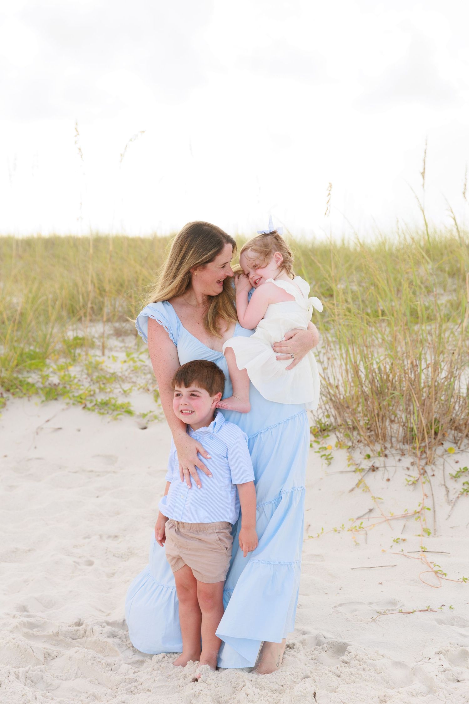 A mother in a blue maxi dress holds her toddler girl while her young son hugs her on a sandy beach.