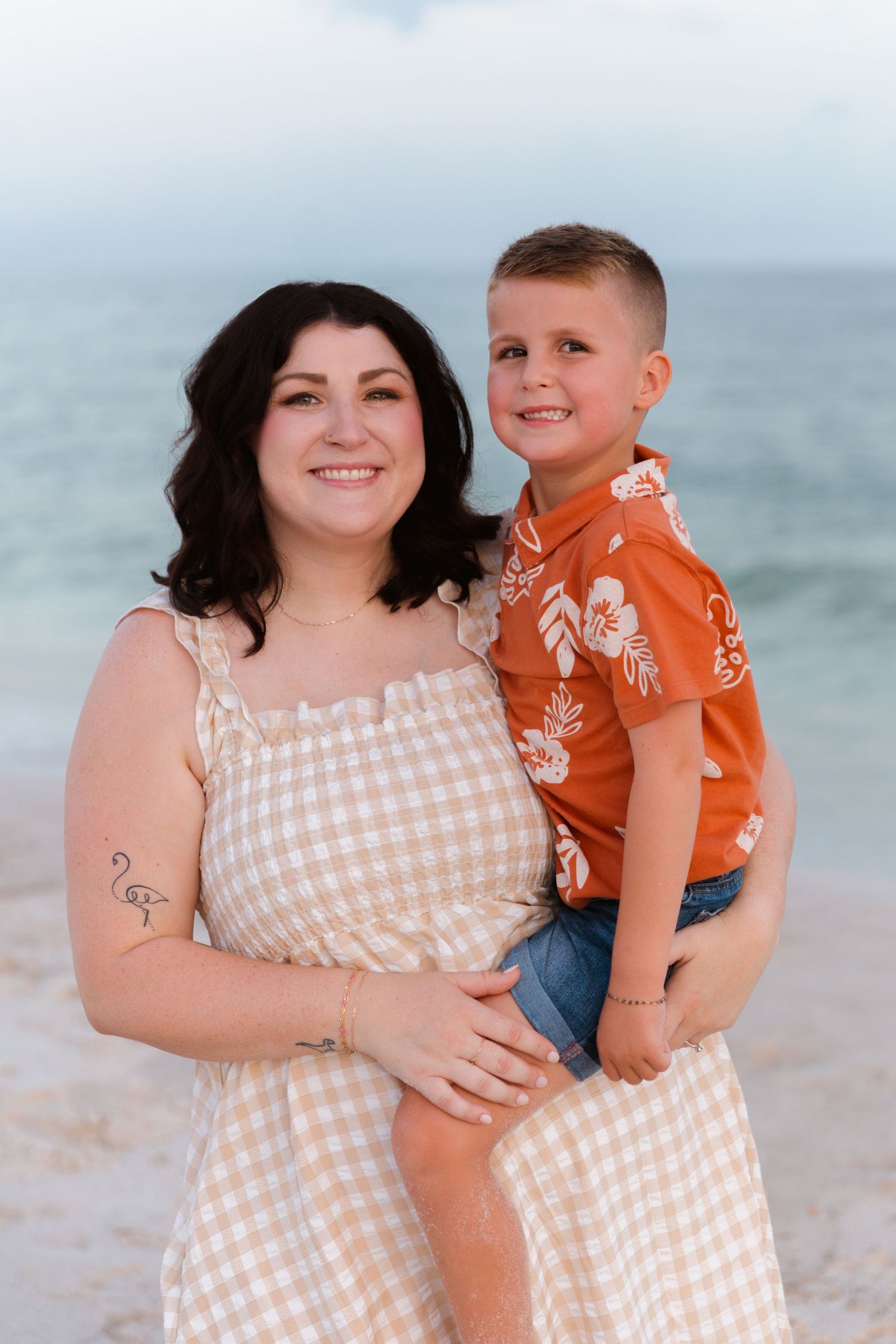 A parent and child wearing coordinating beige gingham and orange outfits pose together on a cloudy beach.