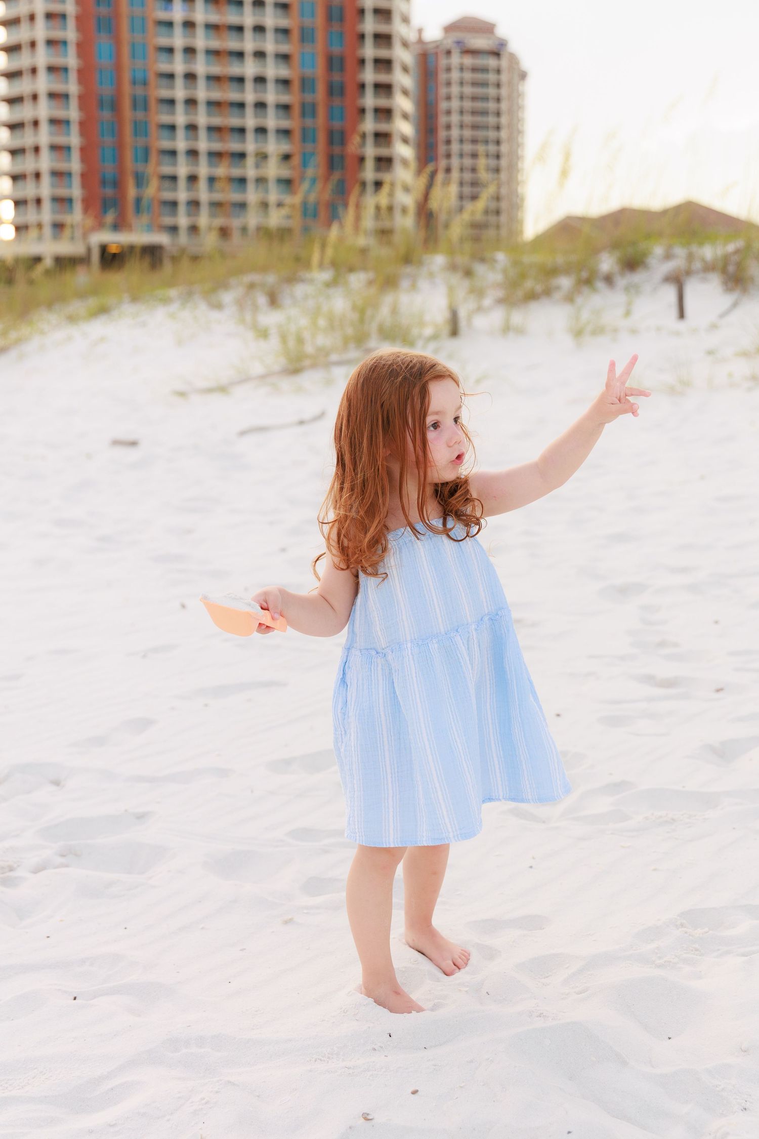 Child in blue dress playing and dancing on white sand beach with condominiums in background.