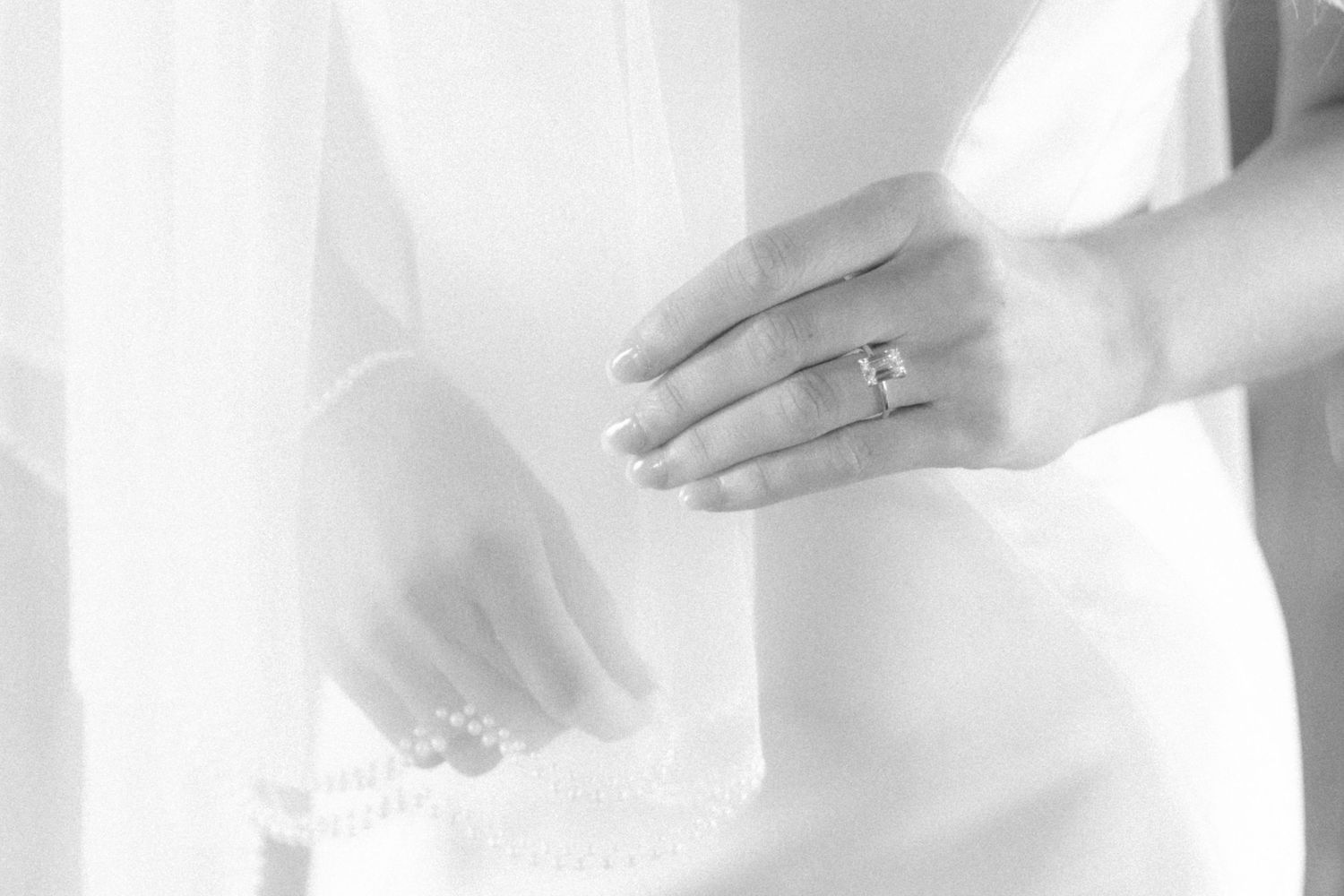 A close-up black and white photo shows hands wearing an elegant engagement ring against a white dress.
