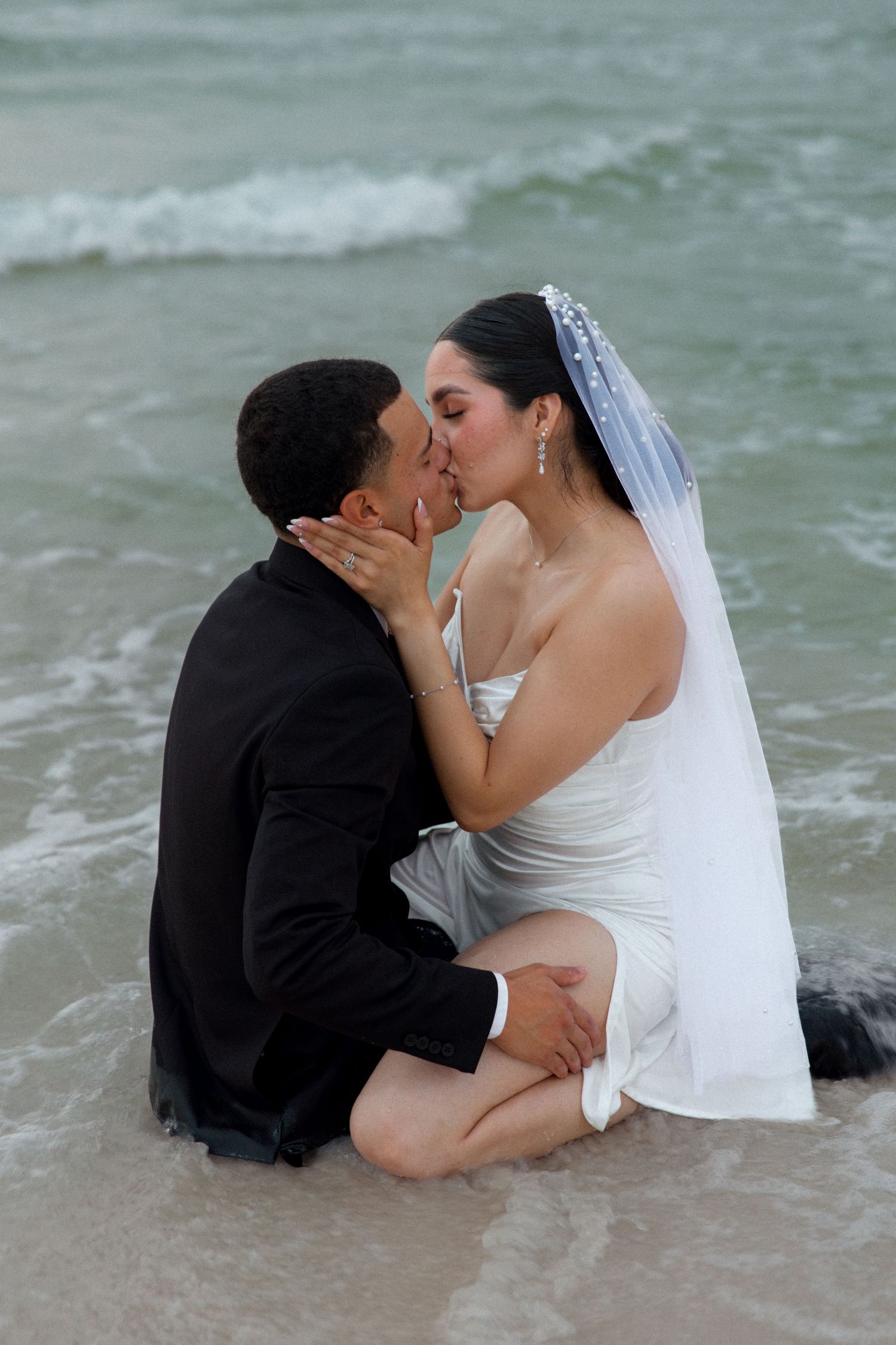Couple shares romantic kiss while sitting on beach near ocean waves.