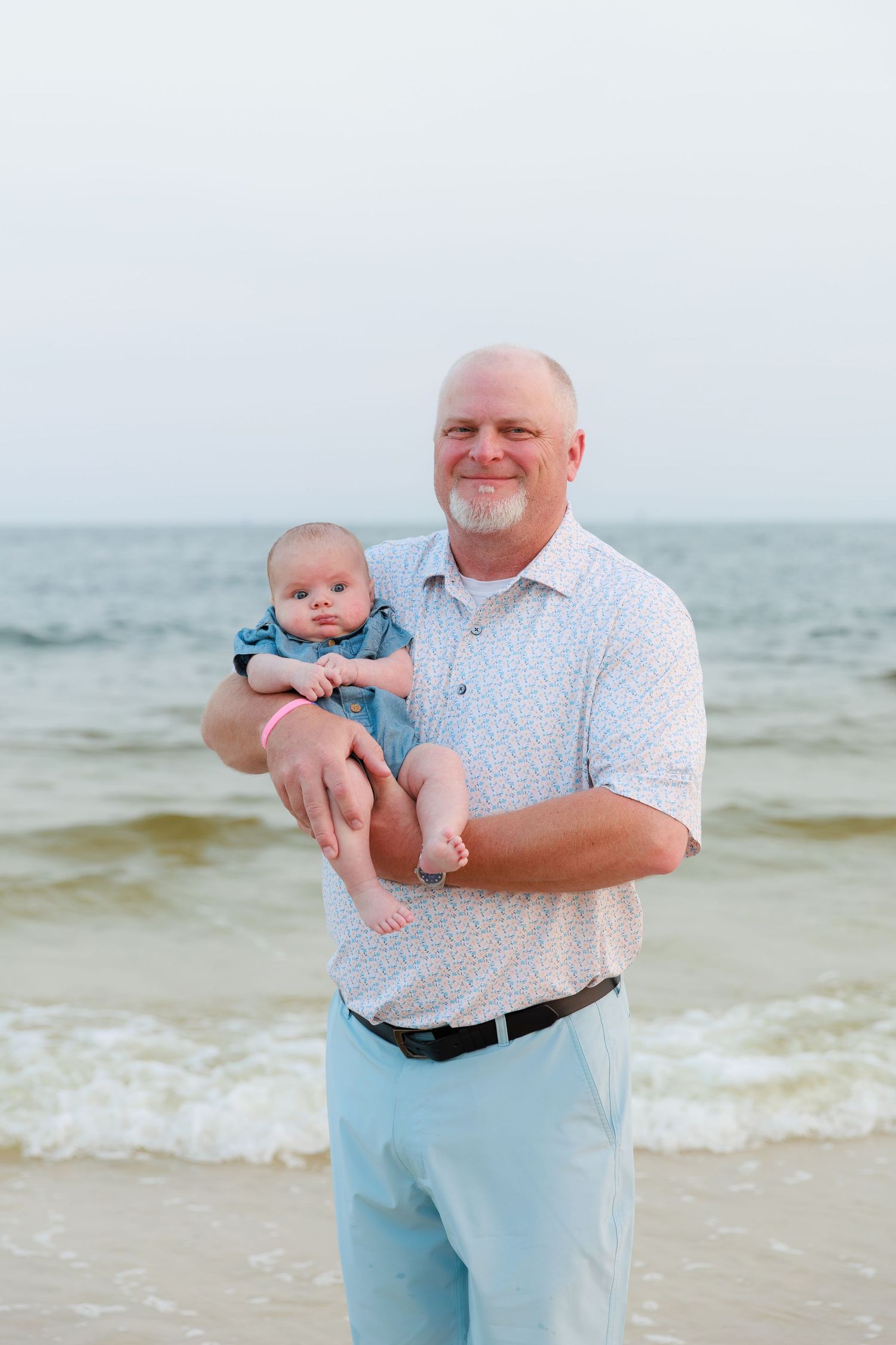 Smiling grandfather holds infant on sunny beach with ocean waves behind them.