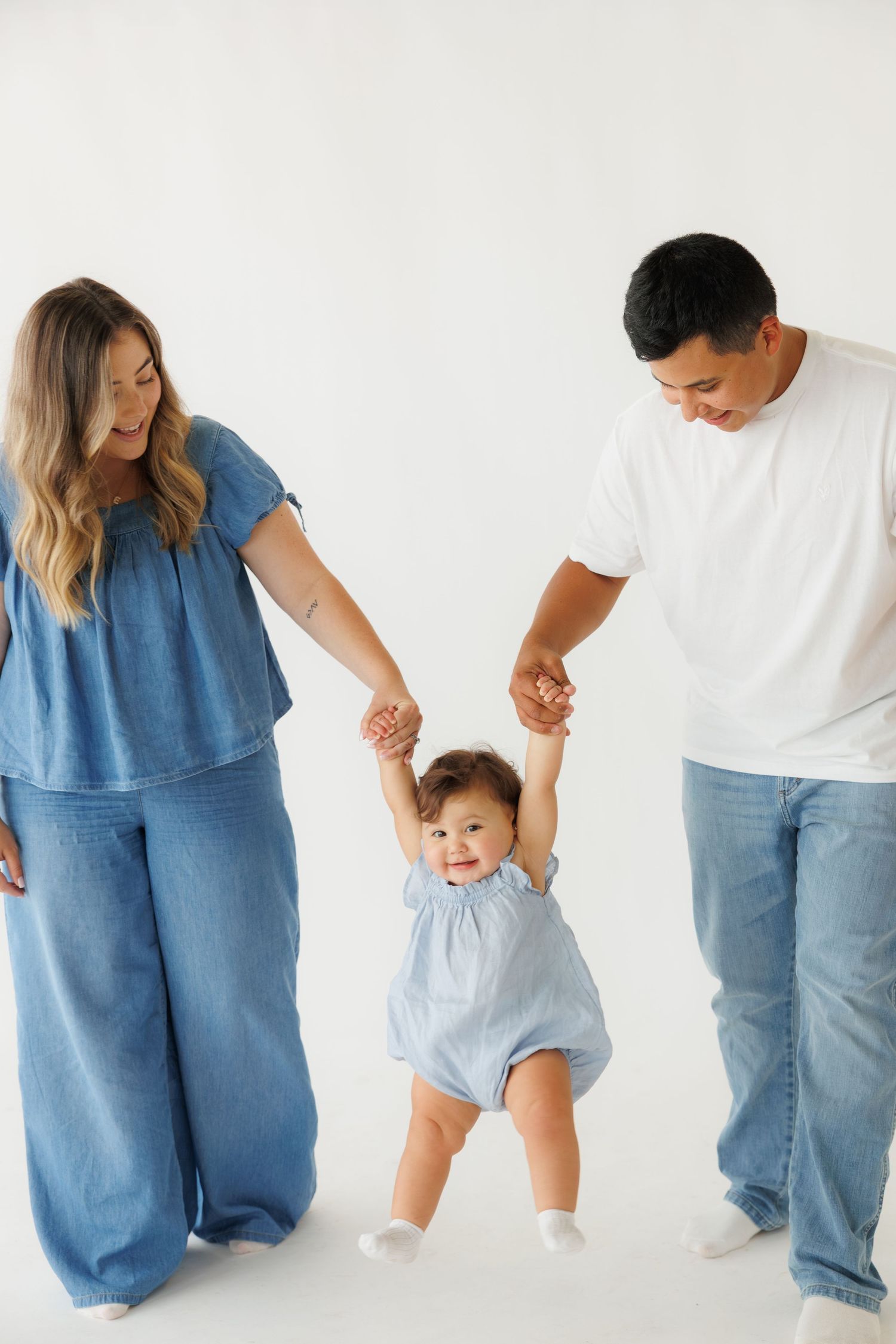 Happy parents in denim helping their baby girl take first steps in white studio setting.