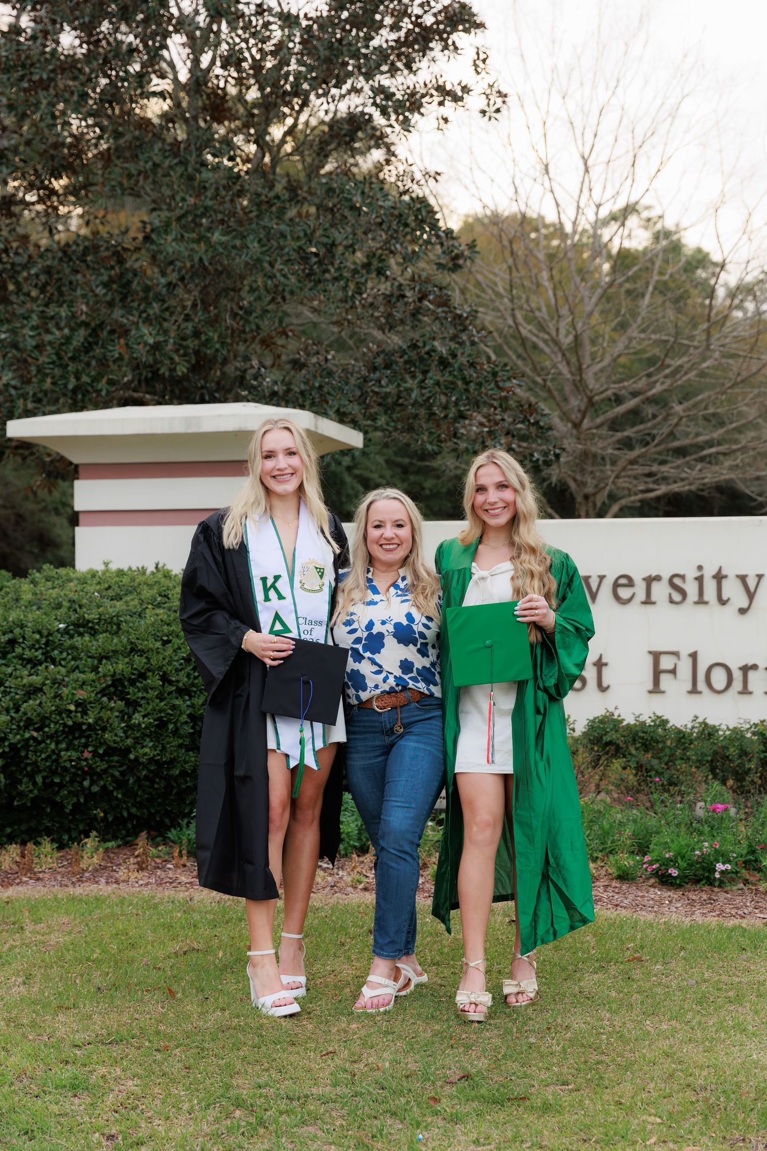 Three women pose in graduation gowns in front of University of West Florida sign.