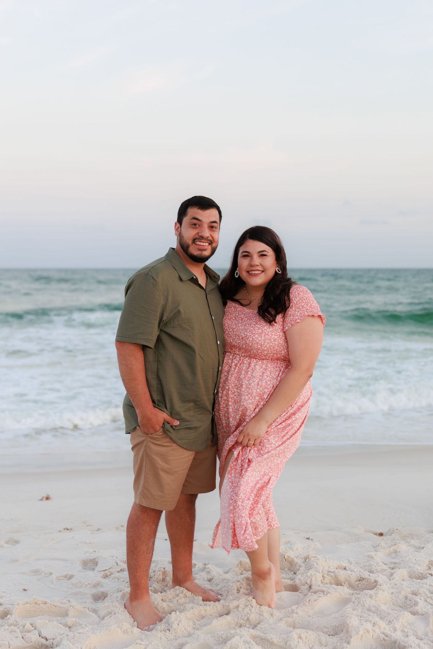 A smiling couple poses barefoot on a sandy beach with ocean waves behind them at dusk.