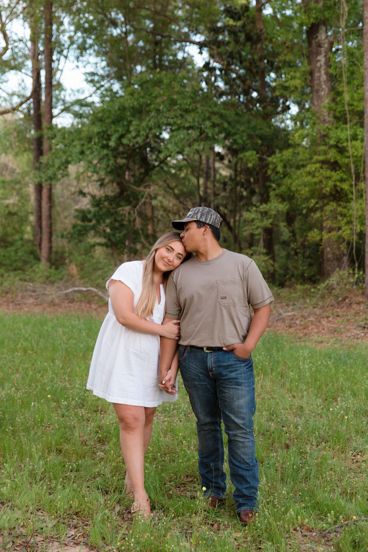 A couple shares a tender kiss in a green meadow, woman in white dress holding hands with man in cap and jeans.