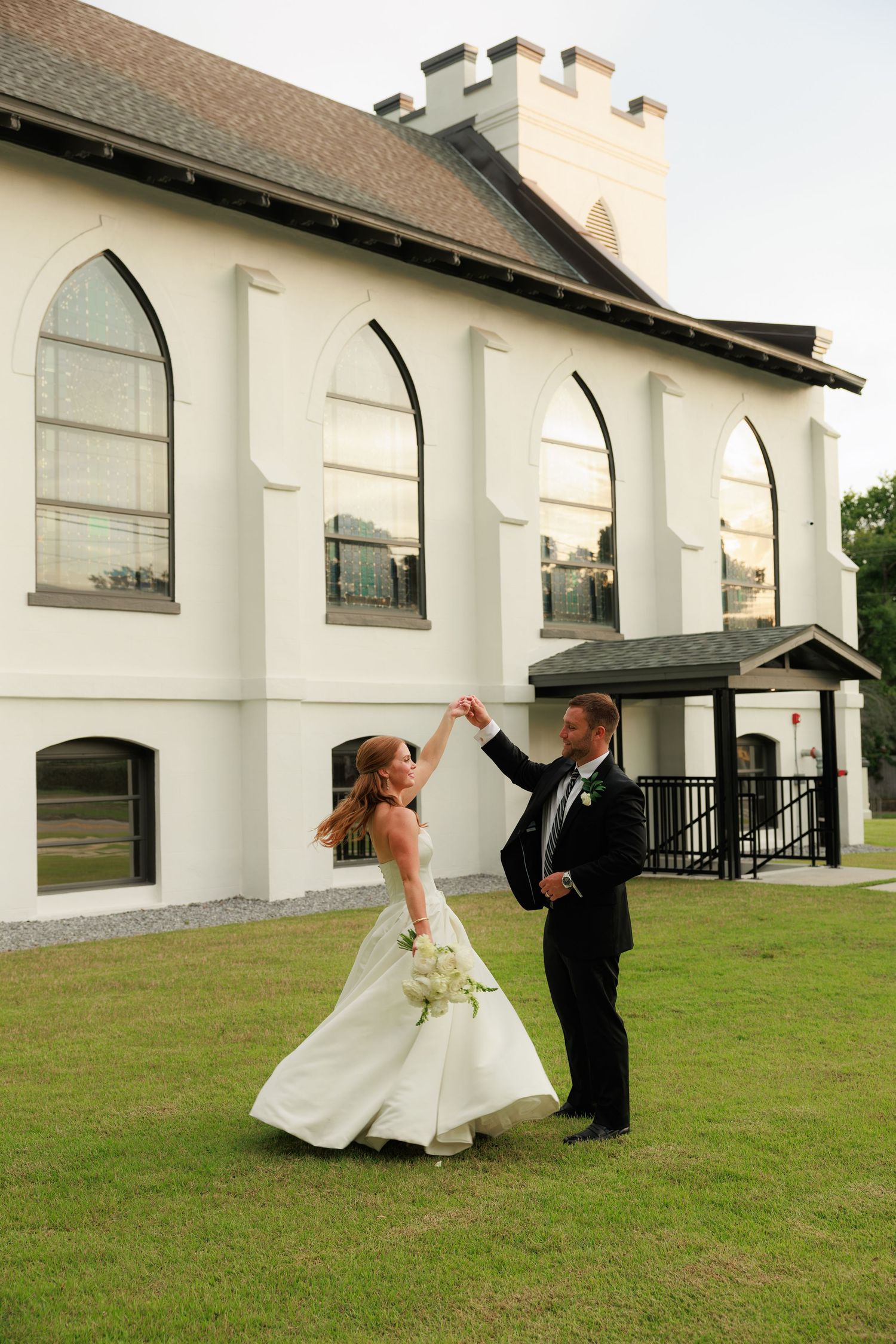 Bride and groom dance joyfully on lawn outside white Gothic church, groom spinning bride in wedding gown.