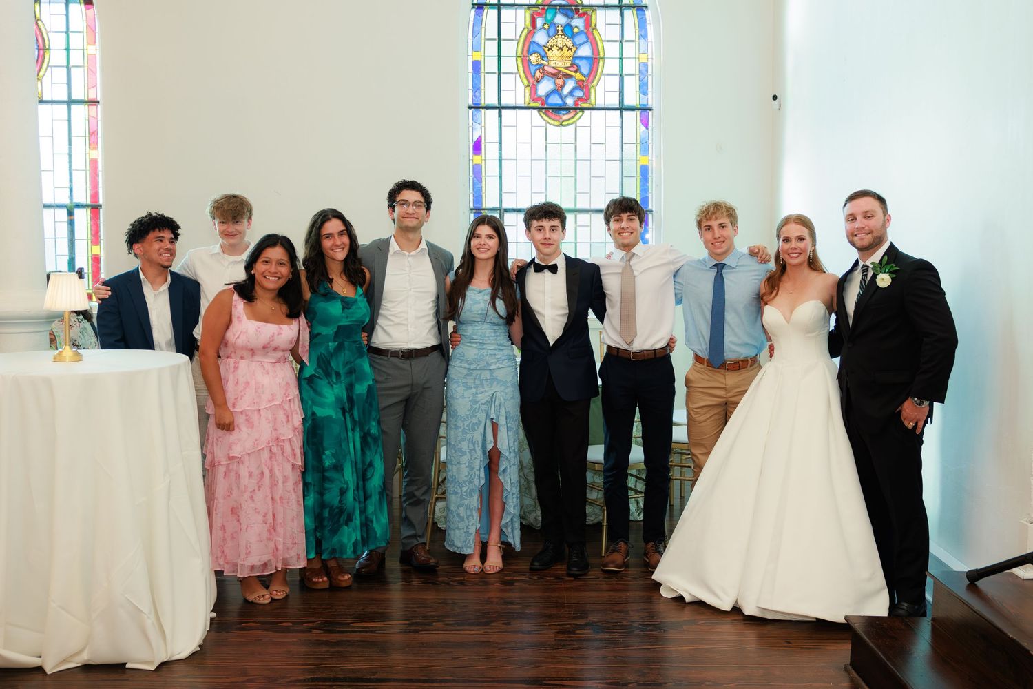 Wedding party poses together in a white chapel with stained glass windows, including bride in white gown and groom in suit.