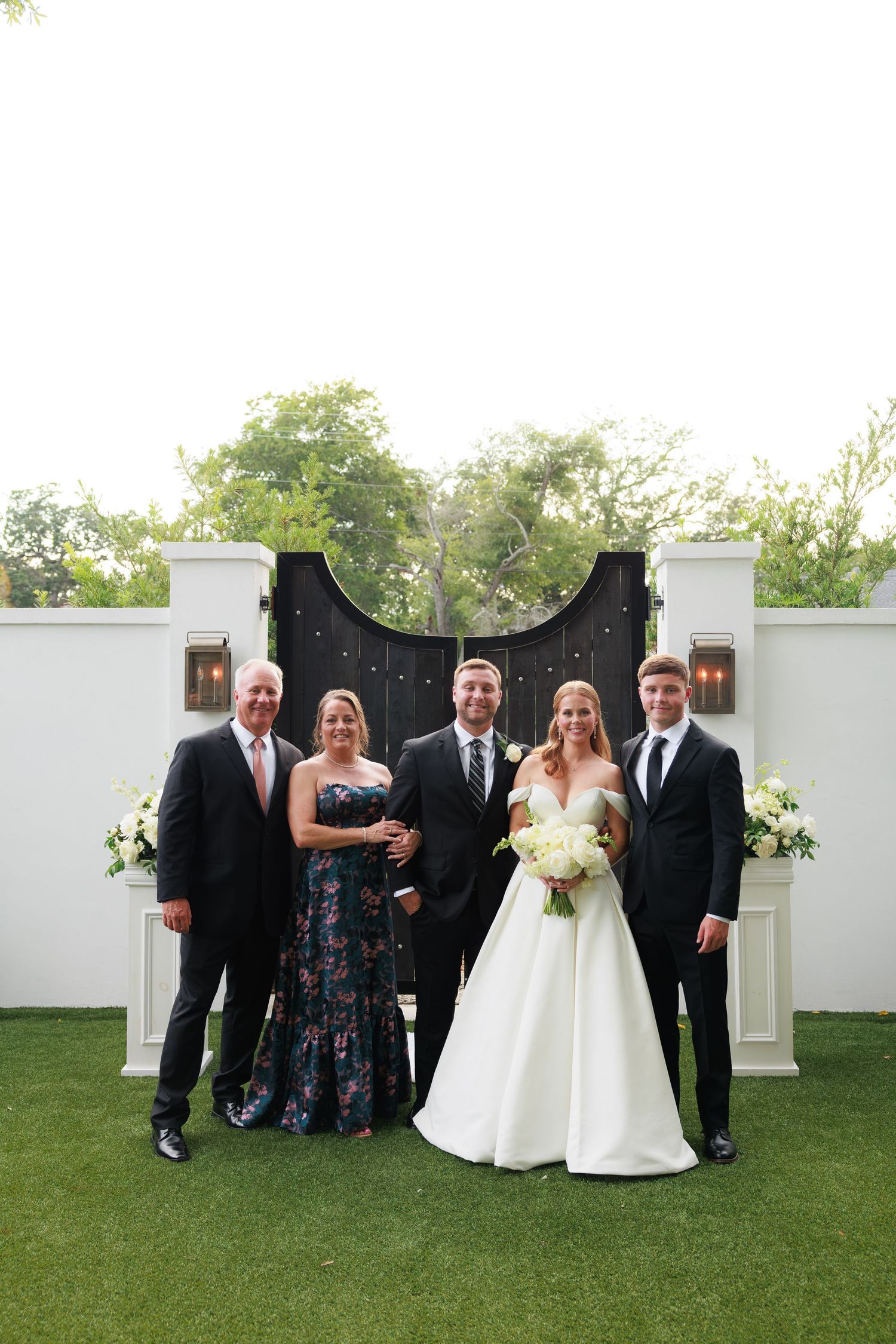 Wedding party of five posing in front of elegant black gates on green lawn, bride in white gown holding bouquet.