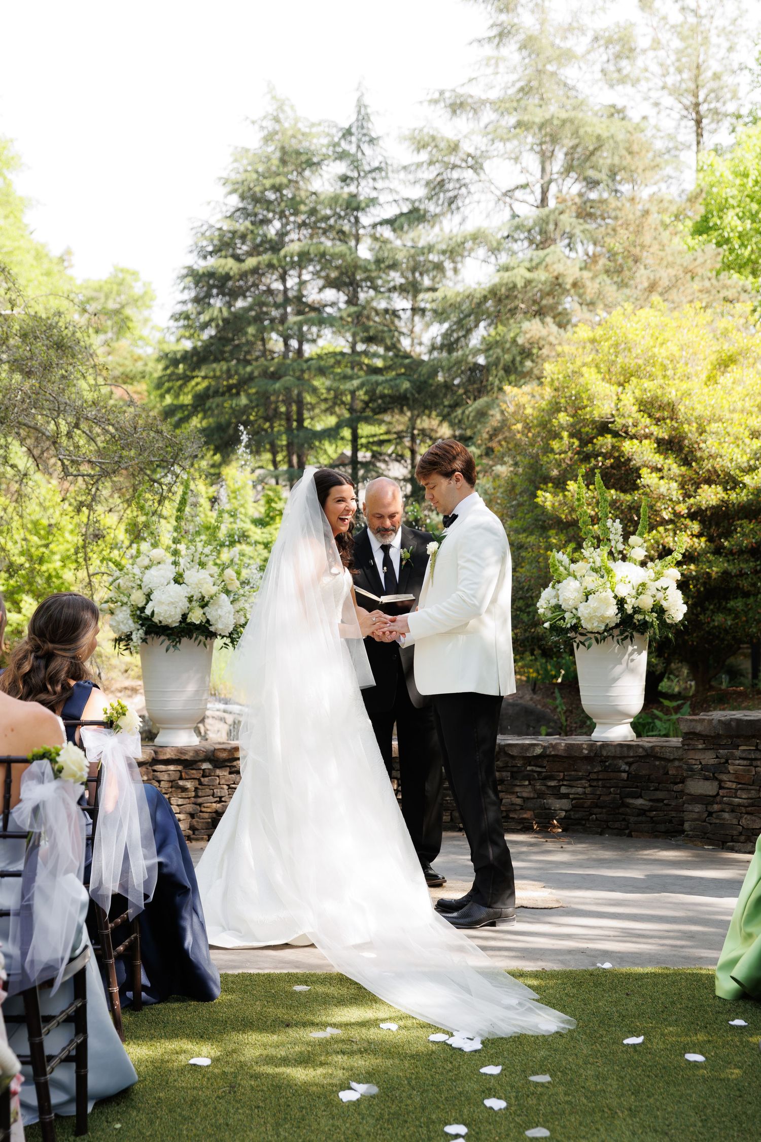 Bride and groom exchange vows outdoors with officiant, surrounded by lush greenery and white floral arrangements.