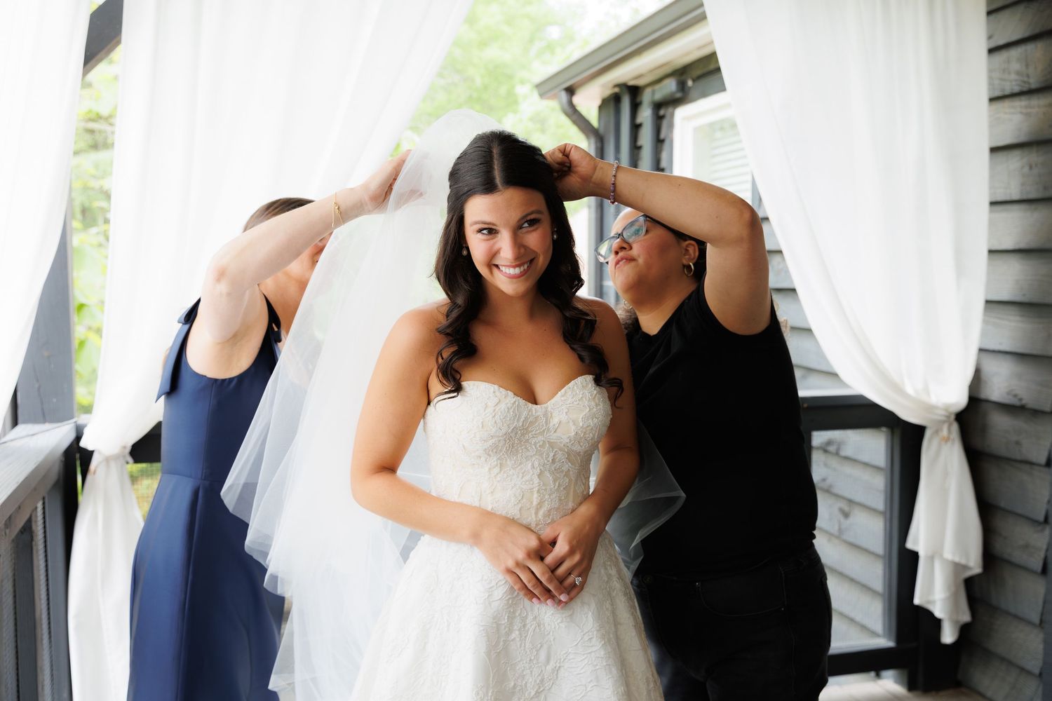 Bride smiling in white strapless gown as bridesmaids adjust her veil on a porch.