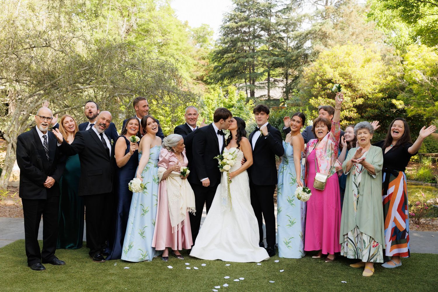 Wedding party posing outdoors in garden, bride and groom centered, guests in colorful formal attire, trees in background.
