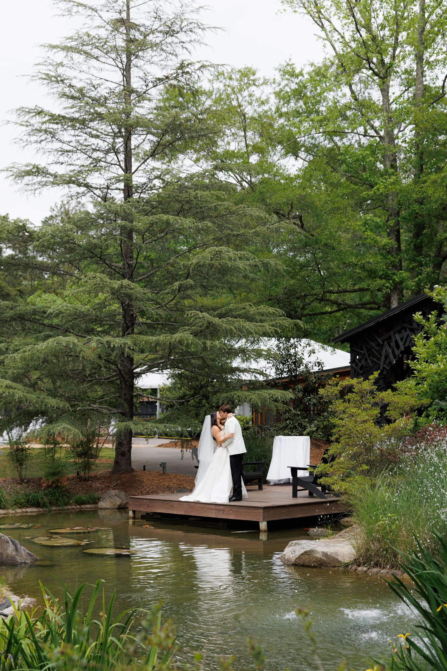 Bride and groom share a kiss on wooden dock surrounded by serene pond and tall trees.
