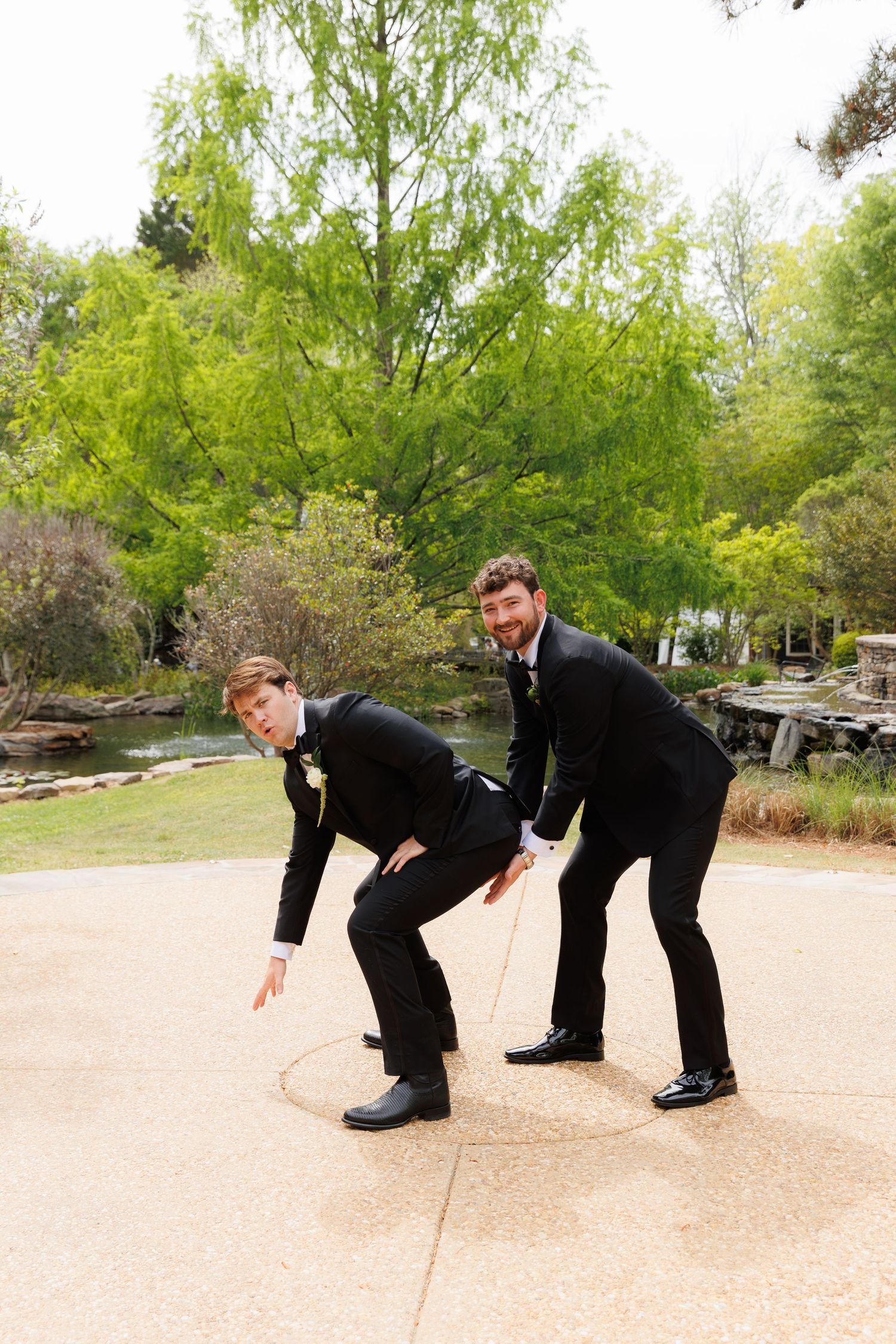Two groomsmen in black tuxedos pose playfully on outdoor patio at wedding venue.