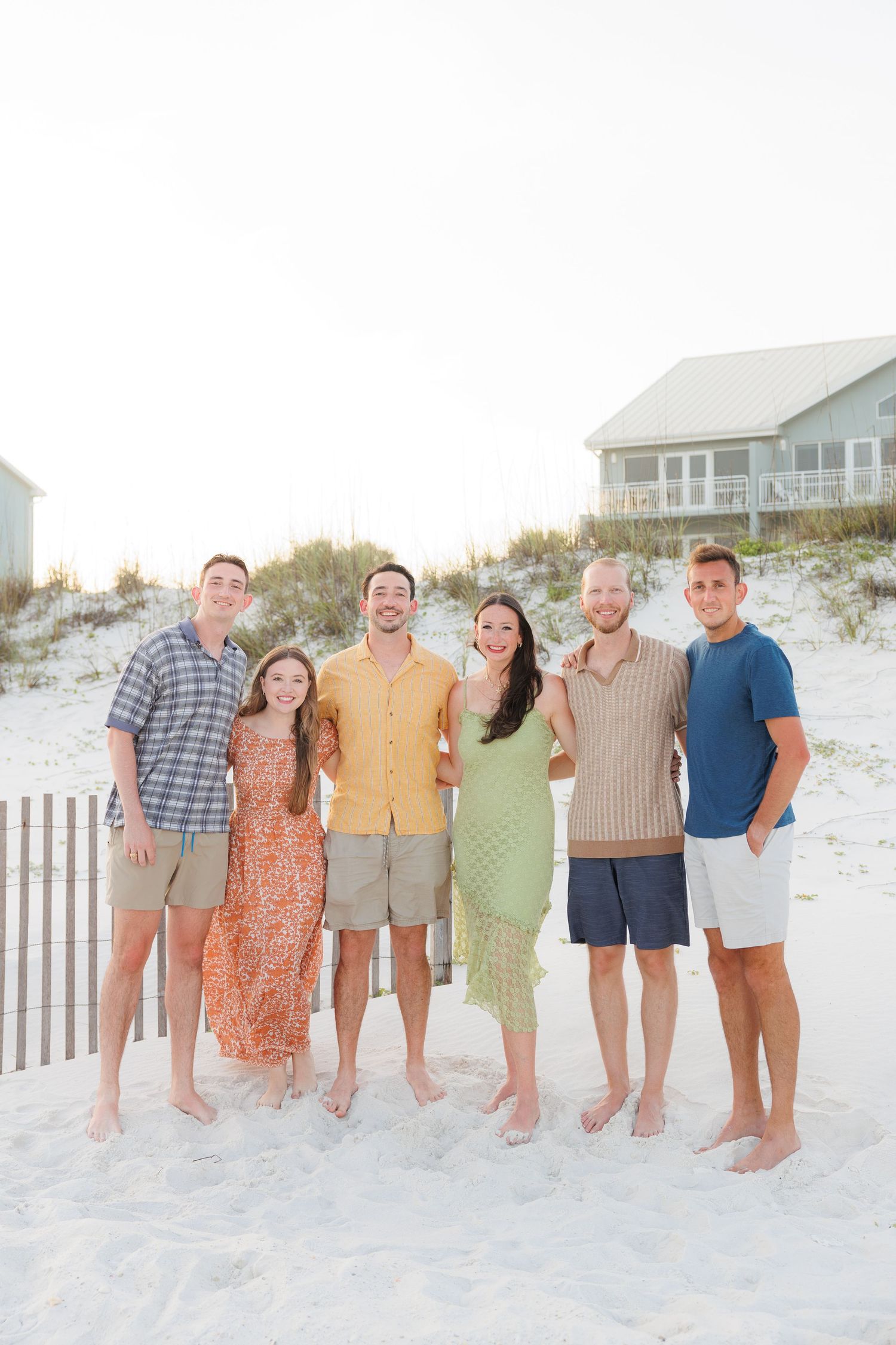 Group of six young adults posing together on sandy beach at sunset.