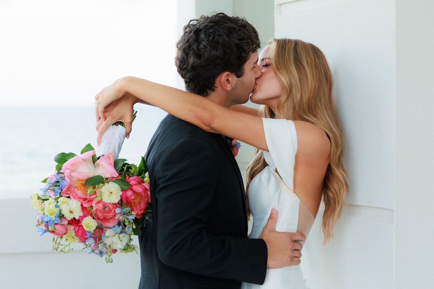 A romantic wedding couple embracing while holding a vibrant bouquet of pink and purple flowers against a white wall.