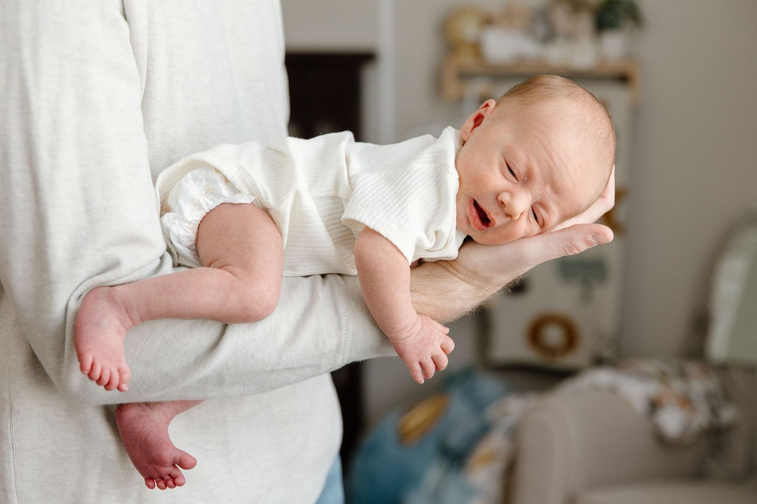 Sleeping baby wearing white onesie rests peacefully on white blanket in home nursery setting.