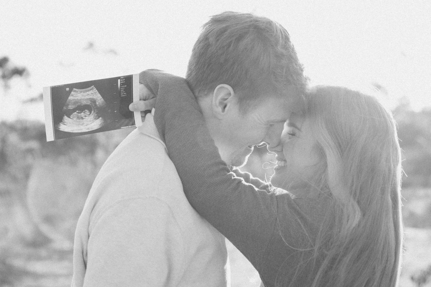 A couple shares an intimate moment while holding up an ultrasound photo in black and white.