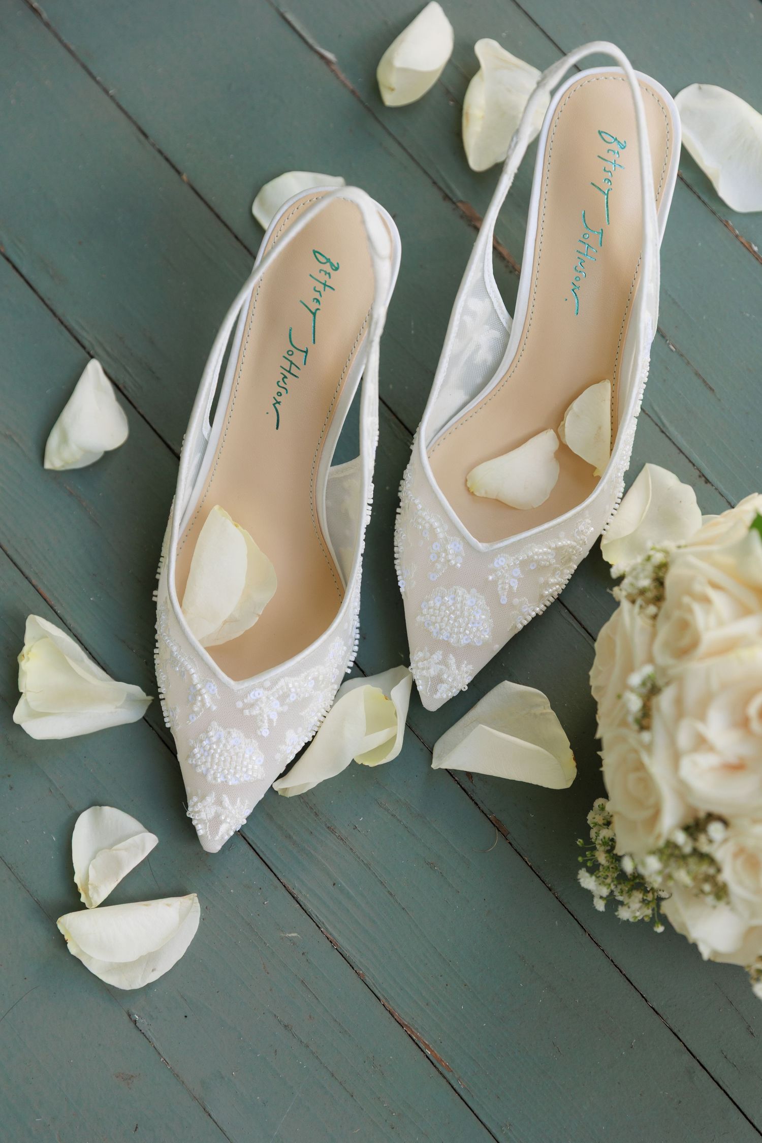 Elegant white lace bridal shoes with delicate beading displayed on a dark green wooden floor next to a white bouquet.