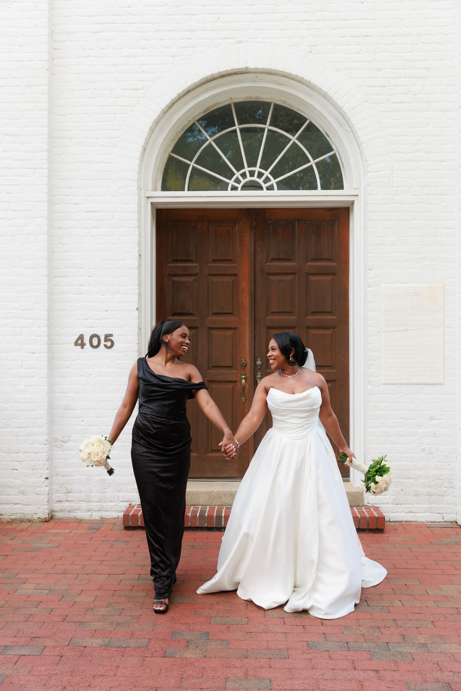 Two people in formal attire holding hands and smiling in front of a historic brown door with arched window at 405.