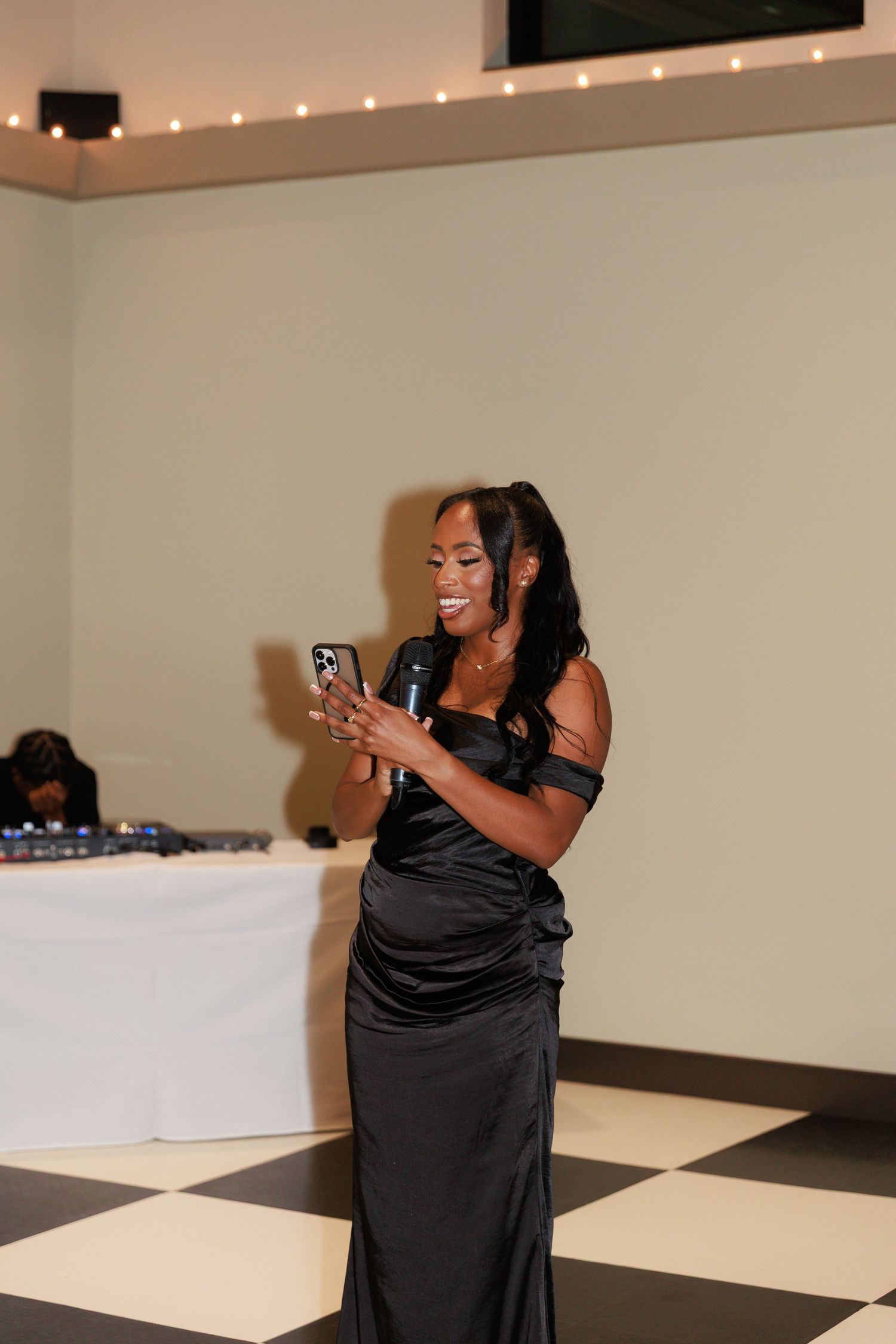 A guest in a black formal dress smiles while looking at their phone during the wedding reception.