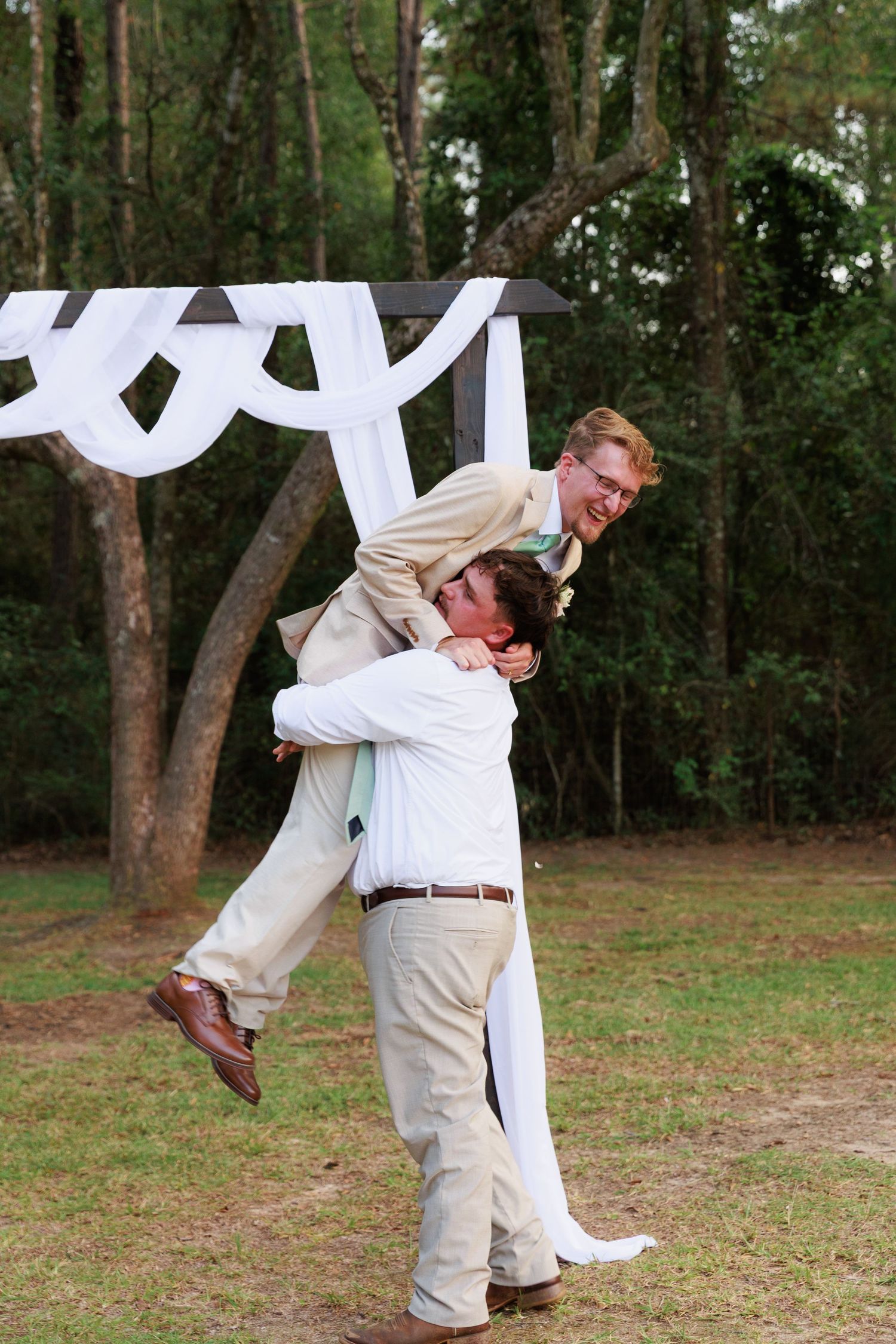Two people in white outfits perform a playful dip pose together in a wooded outdoor setting.
