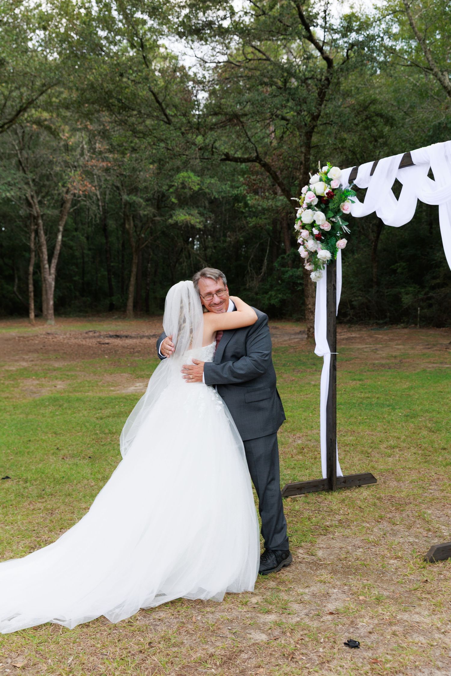 A romantic couple shares a kiss under white fabric decorations in an outdoor wooded setting during their wedding celebration.